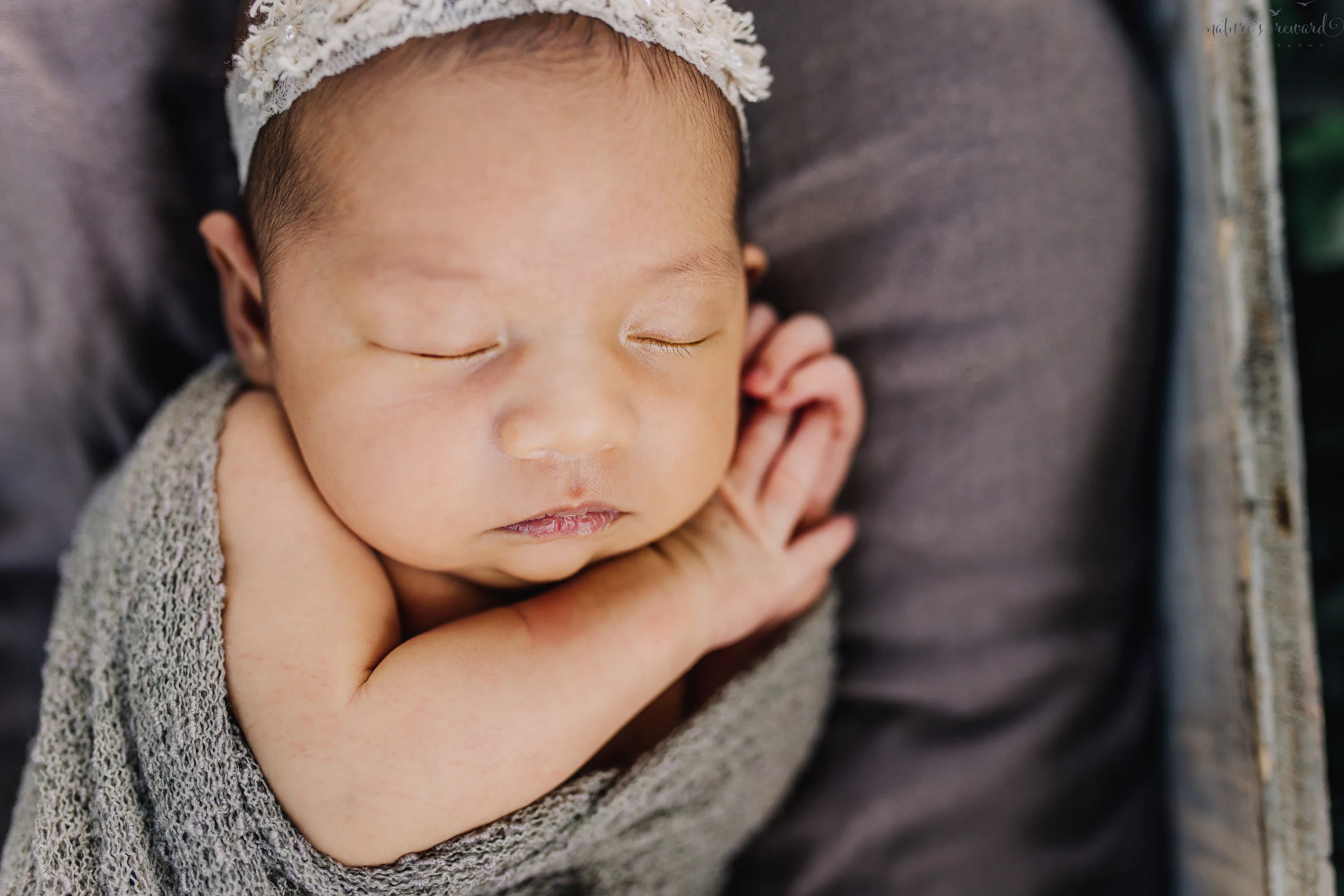 Lovely baby newborn girl swaddled in neutral wearing a white tie back and in a garden surrounded by lush greens in this portrait by Nature’s Reward Photography