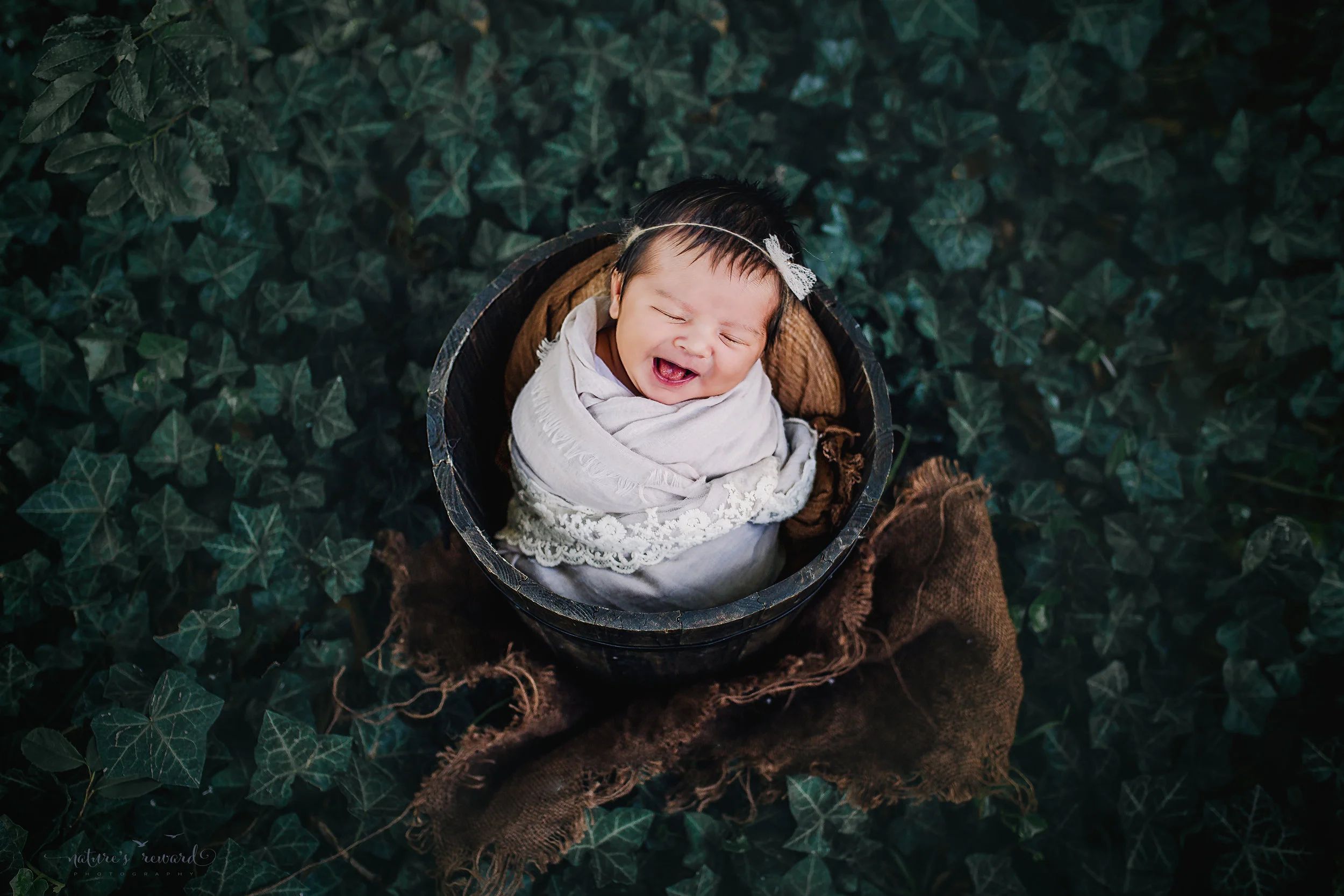 This sweet newborn baby girl in subtle neutral lace adorned wrap in a brown bucket in a bed of ivy smiling. A portrait by Nature’s Reward Photography
