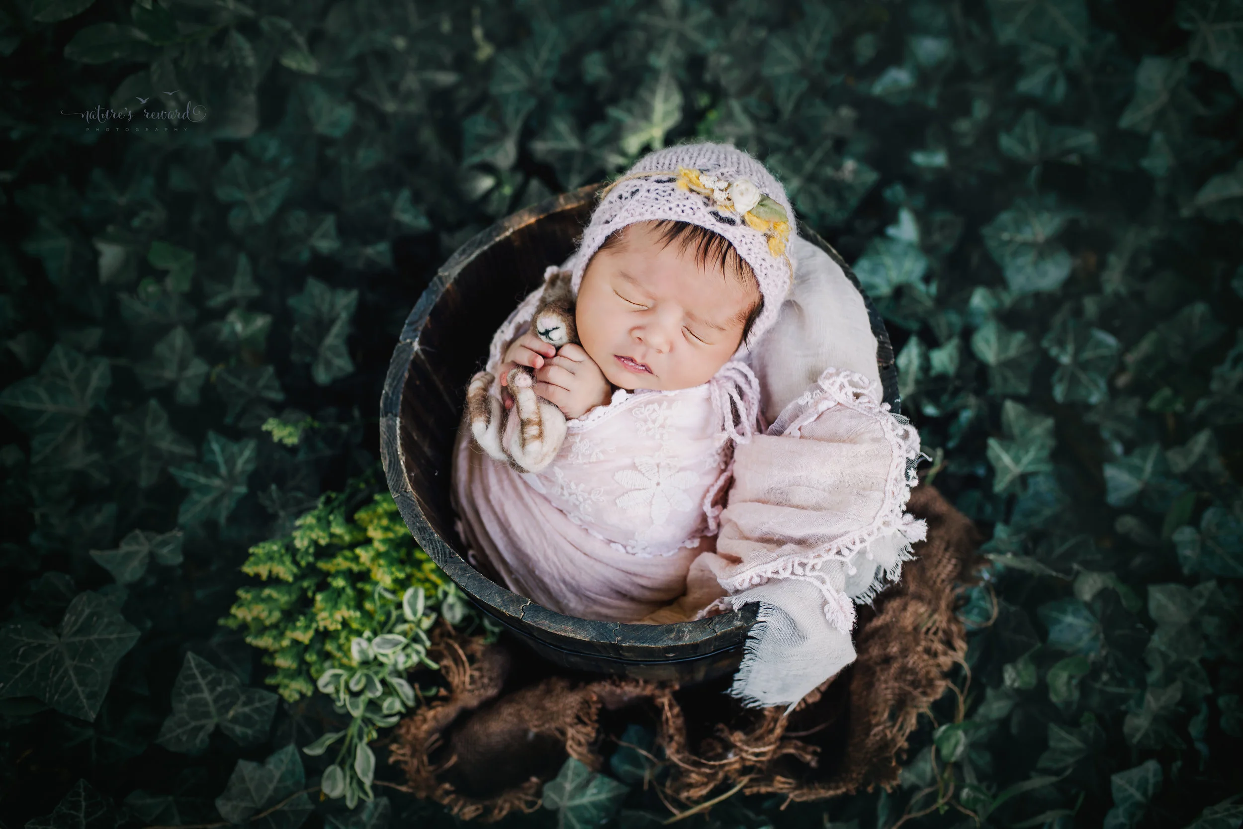 Gorgeous newborn baby girl swaddled in pink wearing a pink bonnet while holding a bunny nested in a bucket in an ivy garden. A portrait by Nature’s Reward Photography