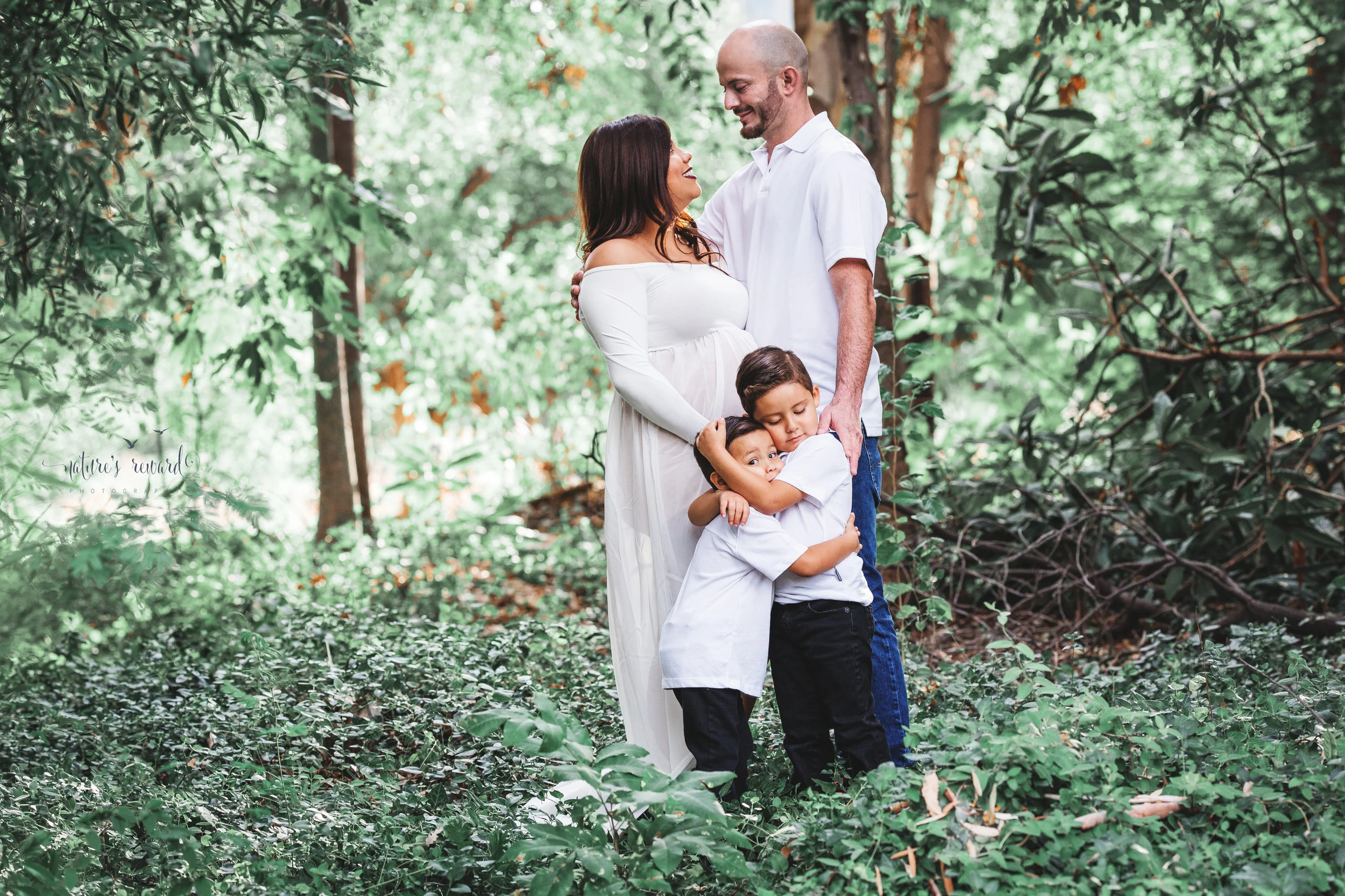 Gorgeous family in whites in a lush green park celebrate the upcoming arrival of their new family member in this lovely maternity session portrait by Nature’s Reward Photography