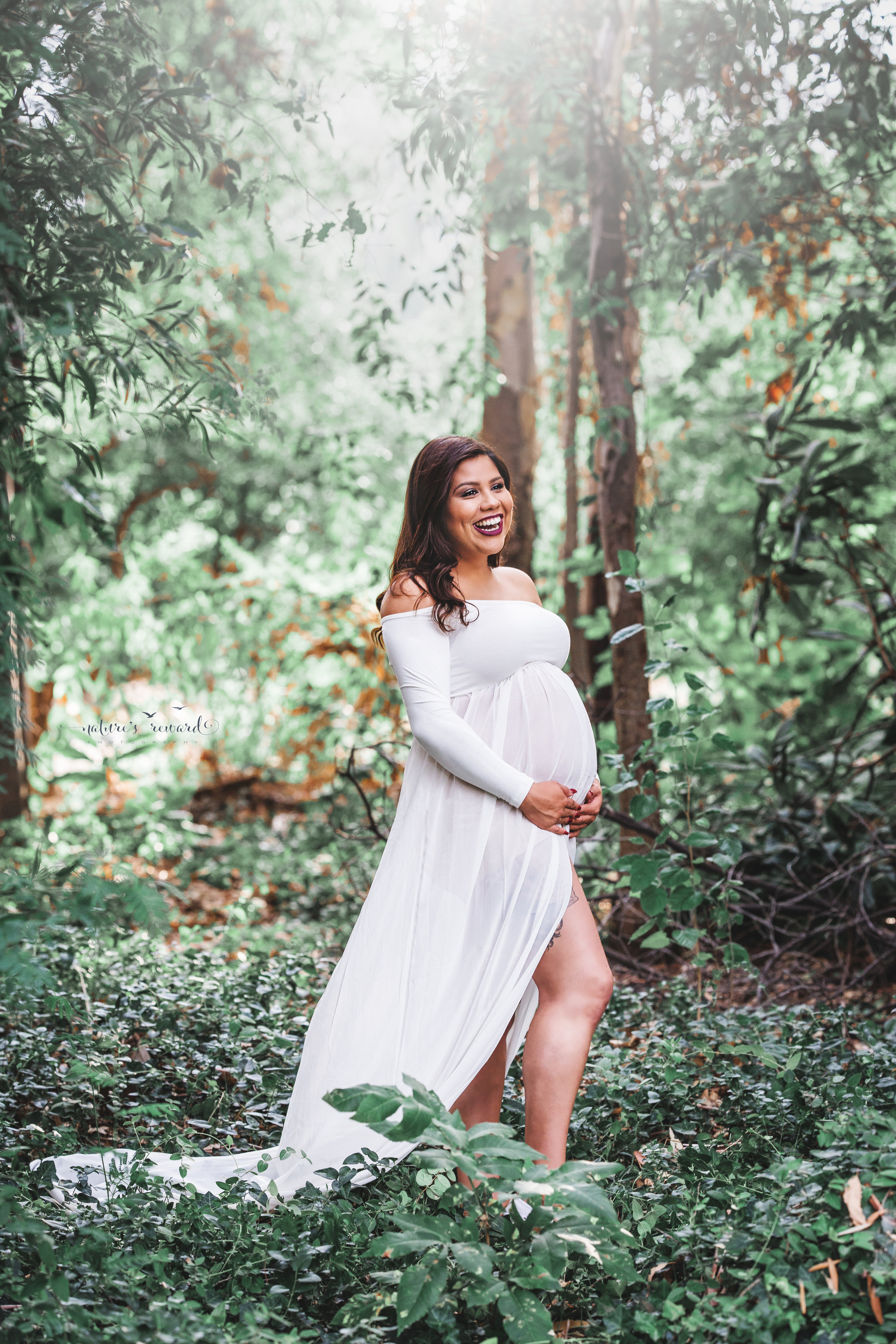 Gorgeous expecting mother in a white maternity gown in. lush green park in this maternity portrait by Nature’s Reward Photography