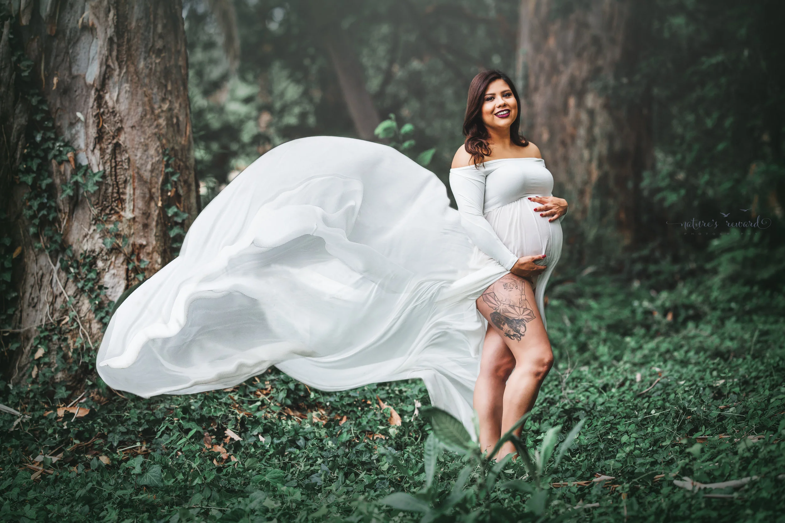 Gorgeous expecting mother in a white maternity gown in. lush green park in this maternity portrait by Nature’s Reward Photography