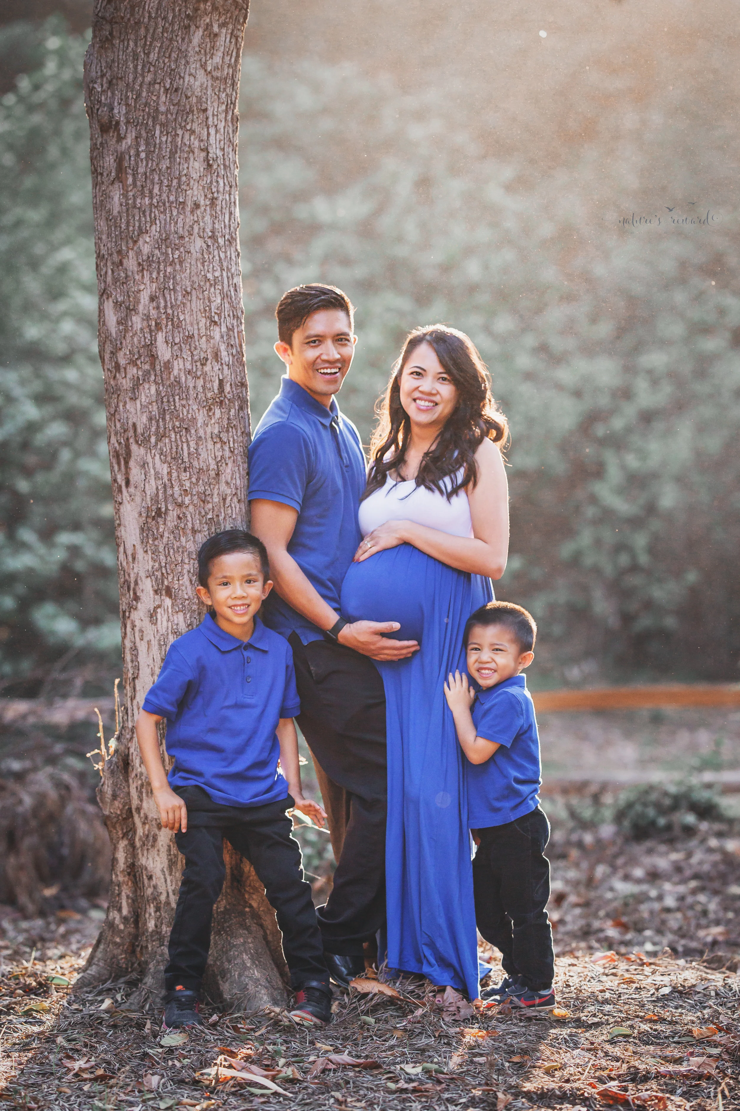 Happy family celebrating the coming of a new baby to complete their family wearing blue to symbolize the gender of the baby in a park setting in this maternity session portrait by Nature’s Reward Photography