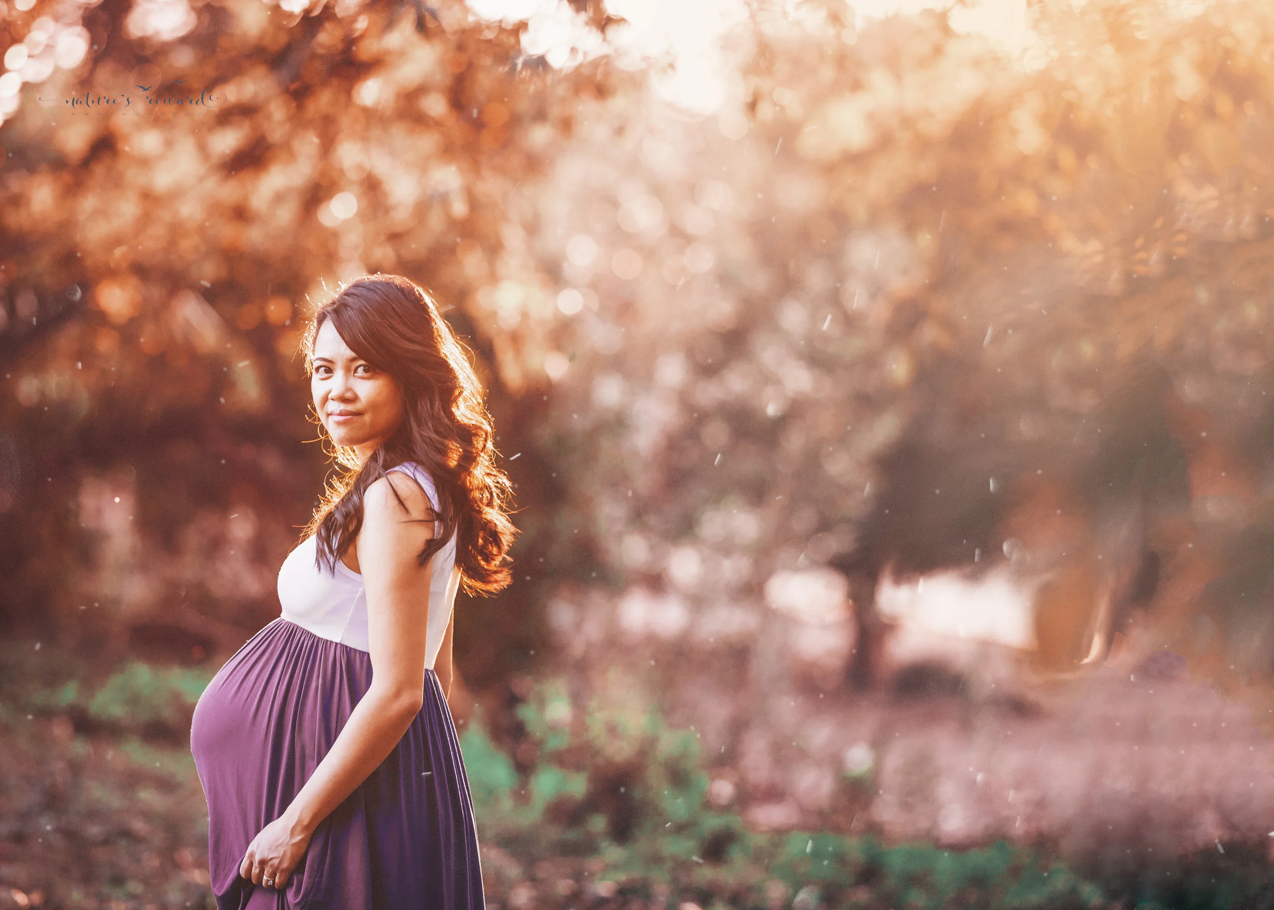 Beautiful full term expecting mother in white and blue dress surrounded by fall colors and warm light in park setting in this maternity session portrait by Nature’s Reward Photography