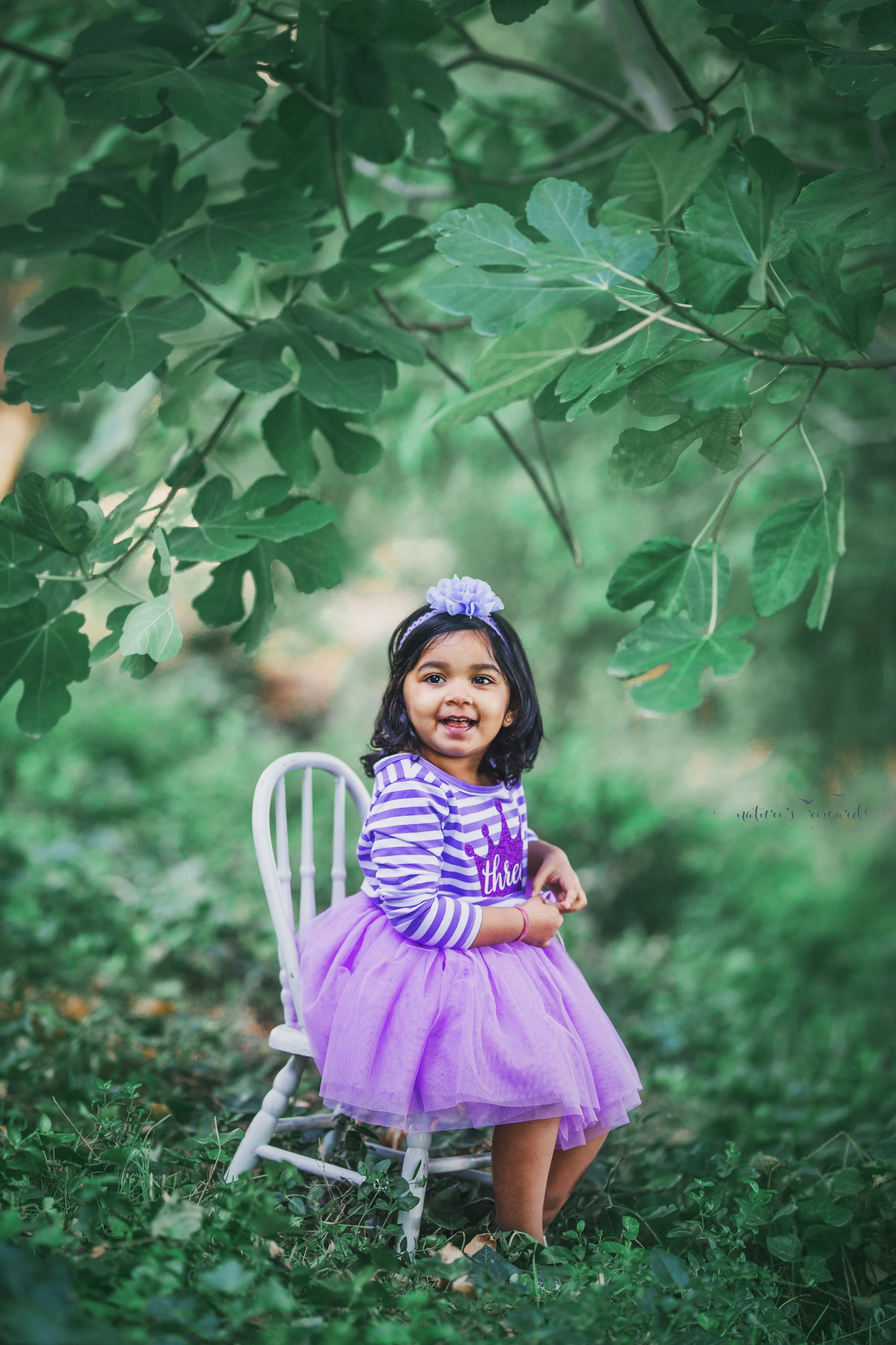 She is 3 birthday photography session while wearing a purple striped and tulle bottom dress sitting on a chair in a magical green park setting a portrait by Nature’s Reward Photography