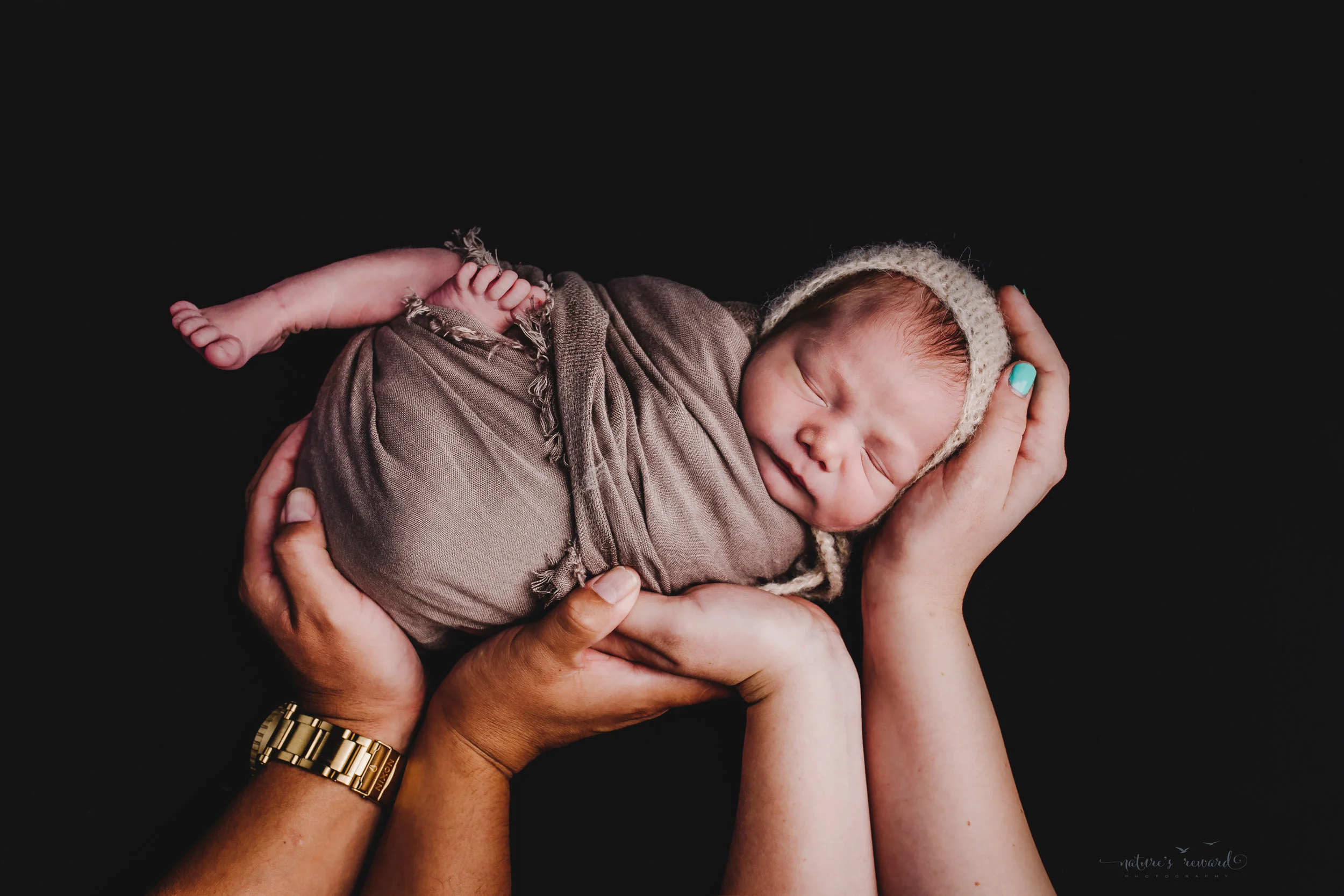 Swaddled and wearing a bonnet being held by his mother’s and father’s hands -a portrait by Nature’s Reward Photography