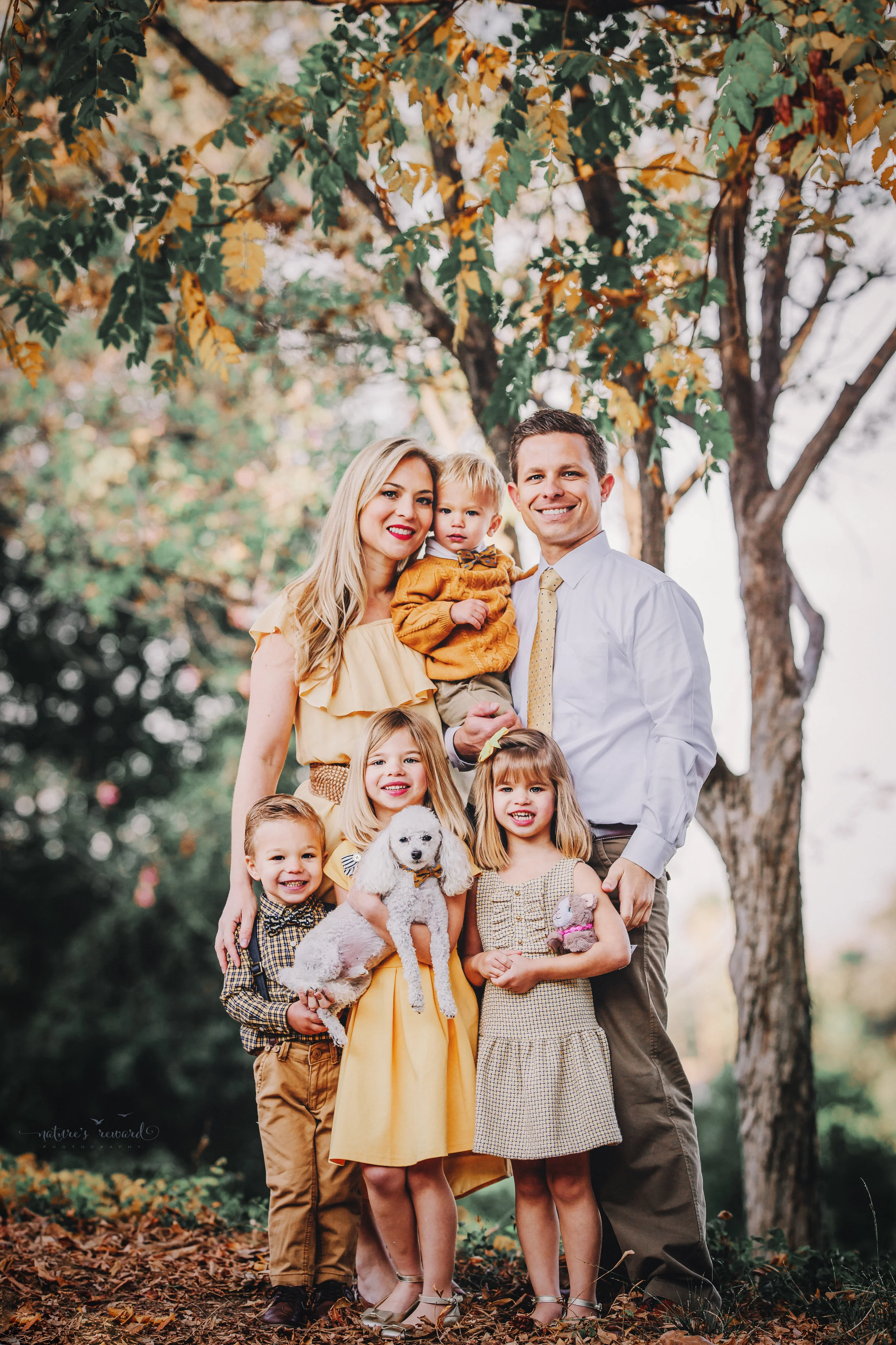 Beautiful family in yellows and tans and whites with their fur baby, 2 daughters and 2 sons under a gorgeous tree with golden leaves - A portrait By Nature’s Reward Photography!