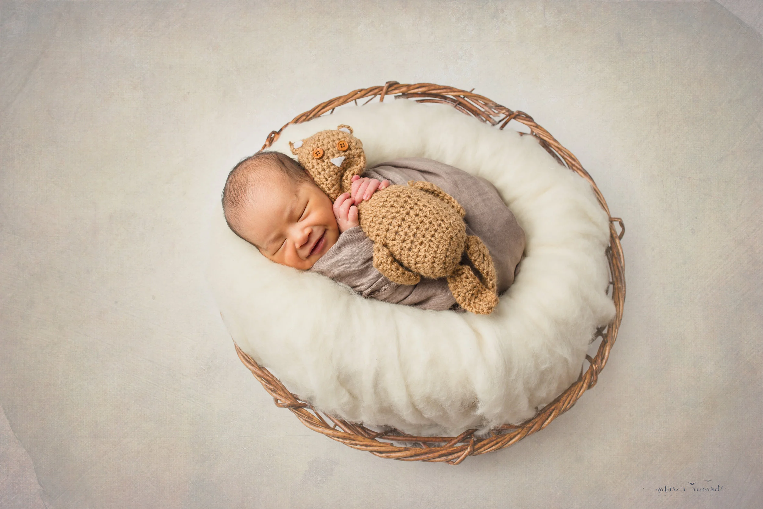 A newborn baby boy in tans and whites, laying a in bowl smiling a portrait by Nature’s Reward Photography