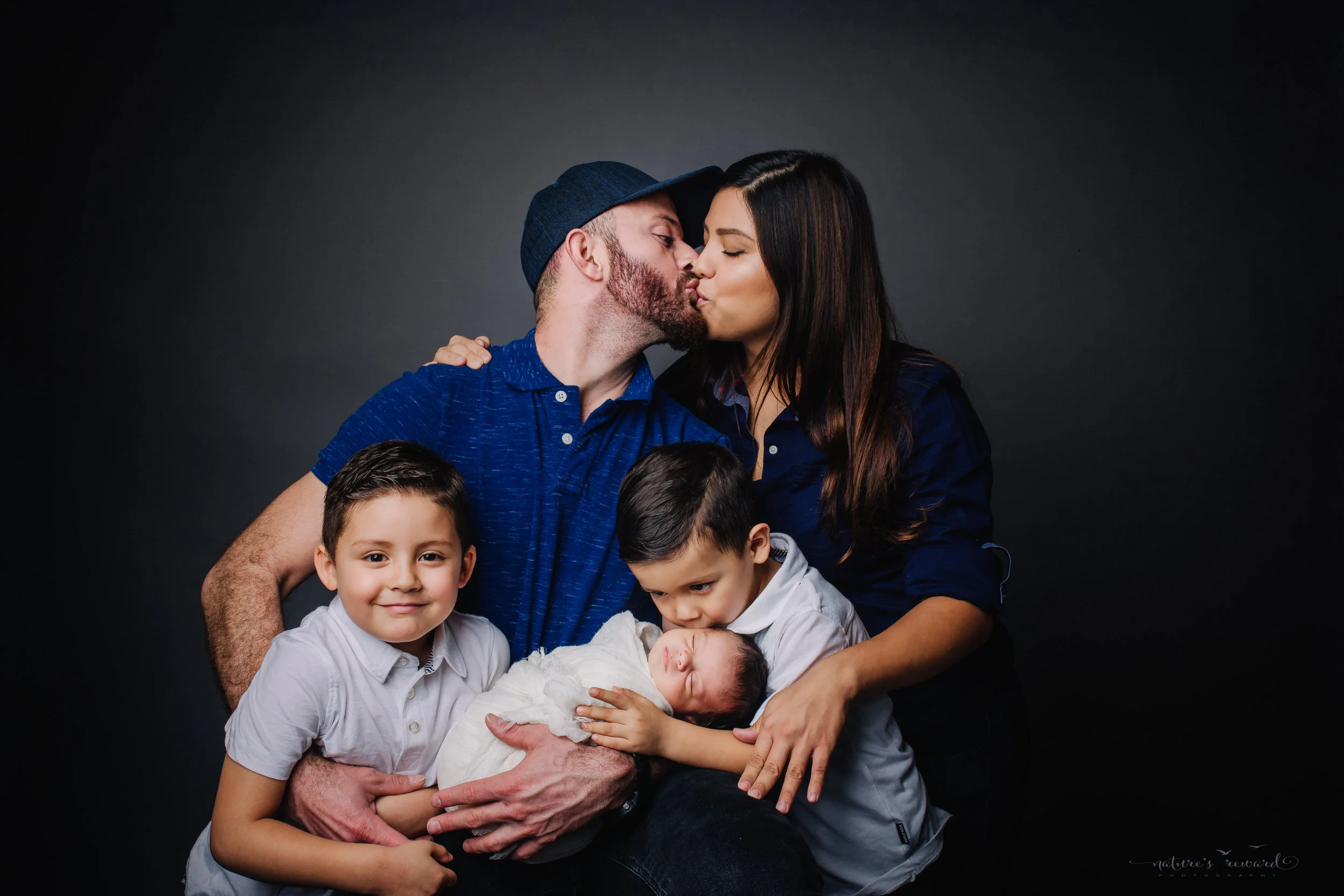 A happy family gives love while holding their newborn baby son swaddled in white - a portrait by Nature’s Reward Photography