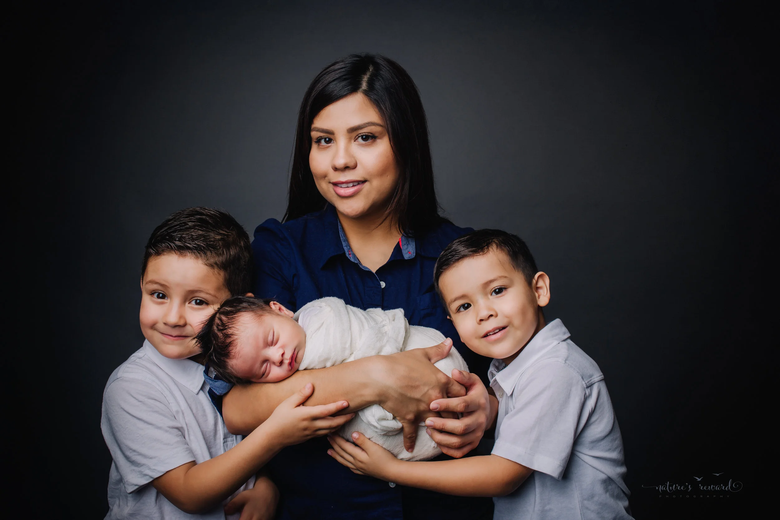 A gorgeous Mother holding her newborn baby boy swaddled in white with her 2 older sons. A family portrait by Nature’s Reward Photography