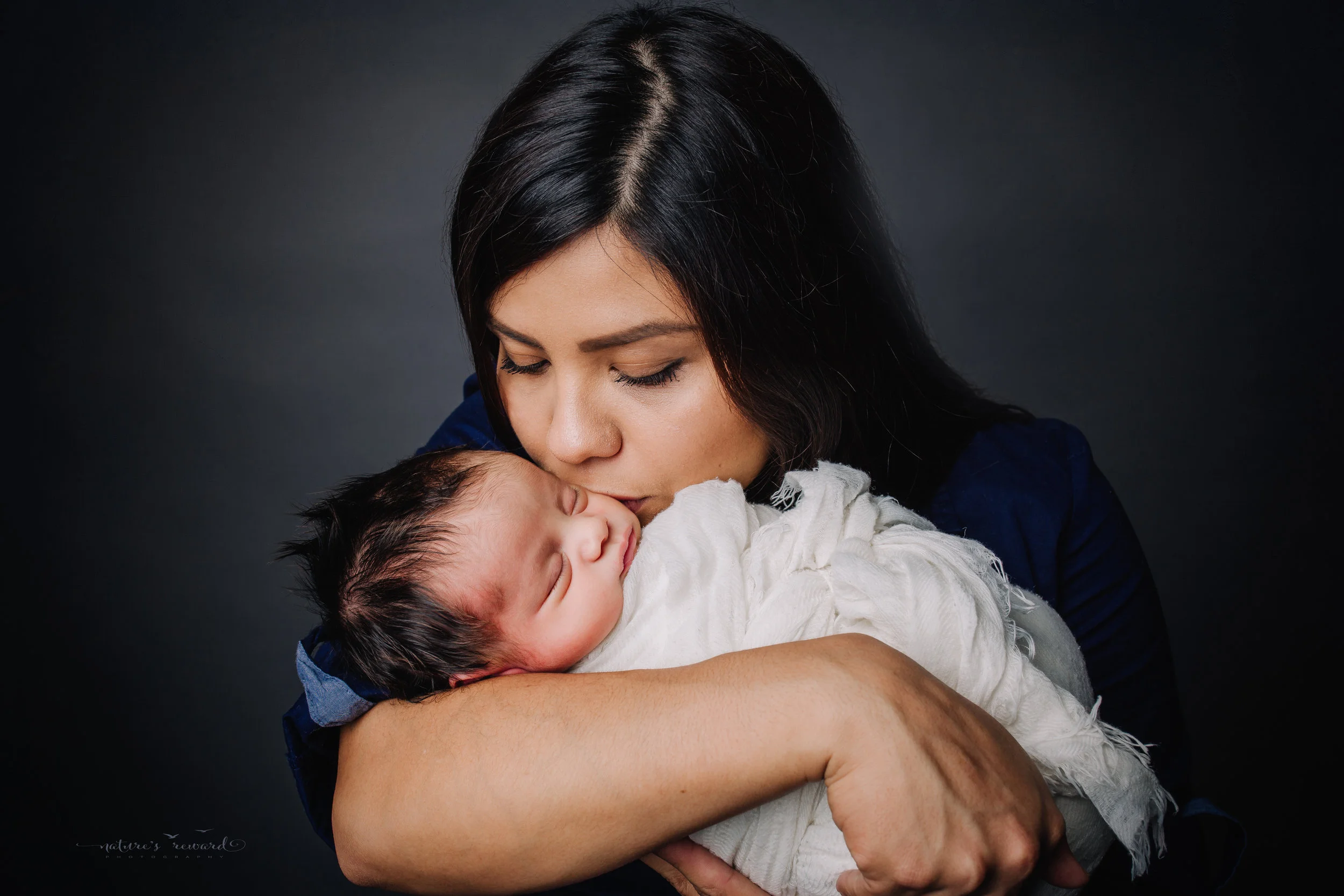 A lovely mother kisses er newborn baby boy swaddled in white - a portrait by Nature’s Reward Photography