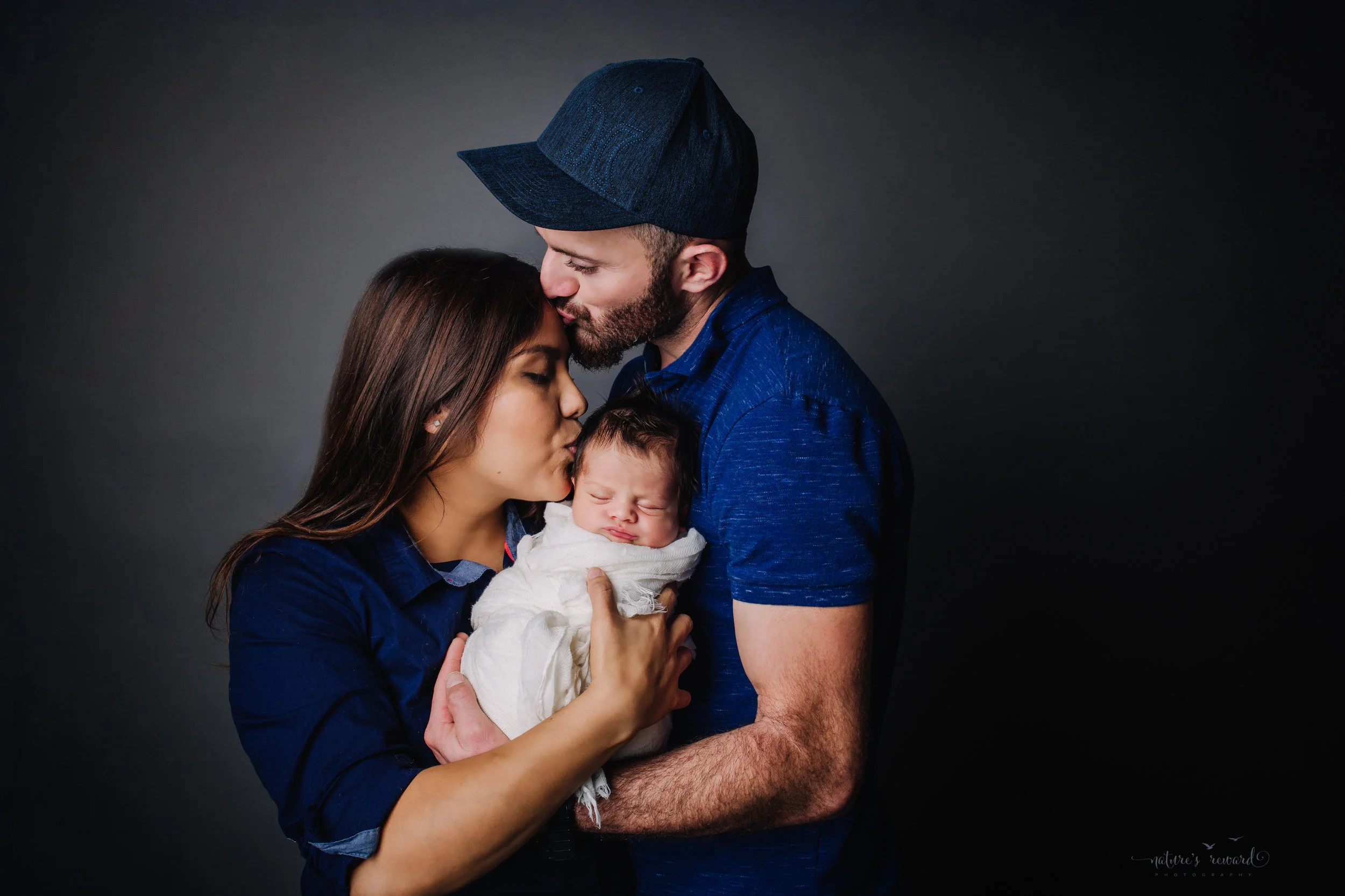 A newborn baby boy swaddled in white in his parent’s arms - a portrait by Nature’s Reward Photography