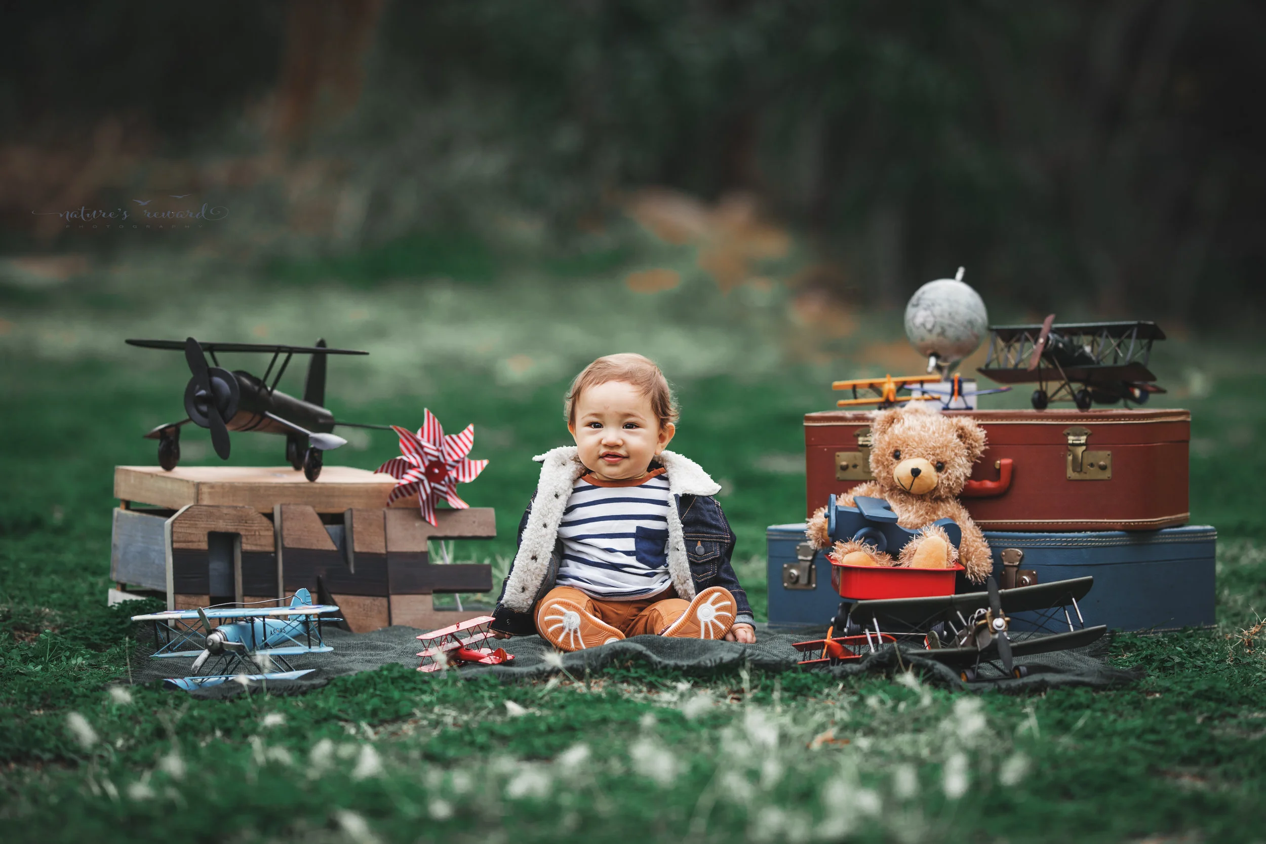Beautiful baby boy in tans and a white and blue striped shirt and jean jacket in his airplane themed cake smash set in this portrait by Nature’s Reward photography