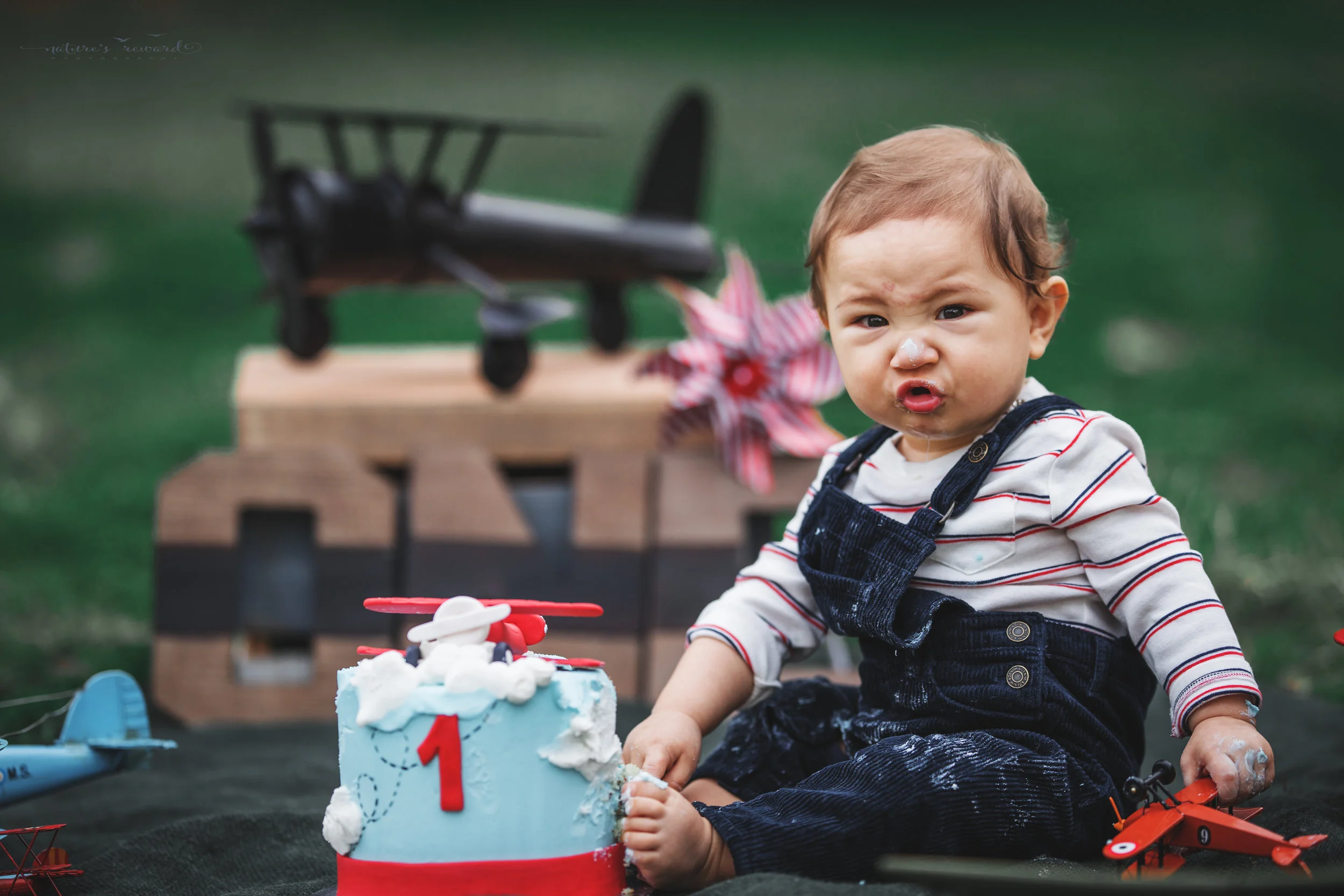 Beautiful baby boy in tans and a white and blue striped shirt and jean jacket in his airplane themed cake smash set in this portrait by Nature’s Reward photography