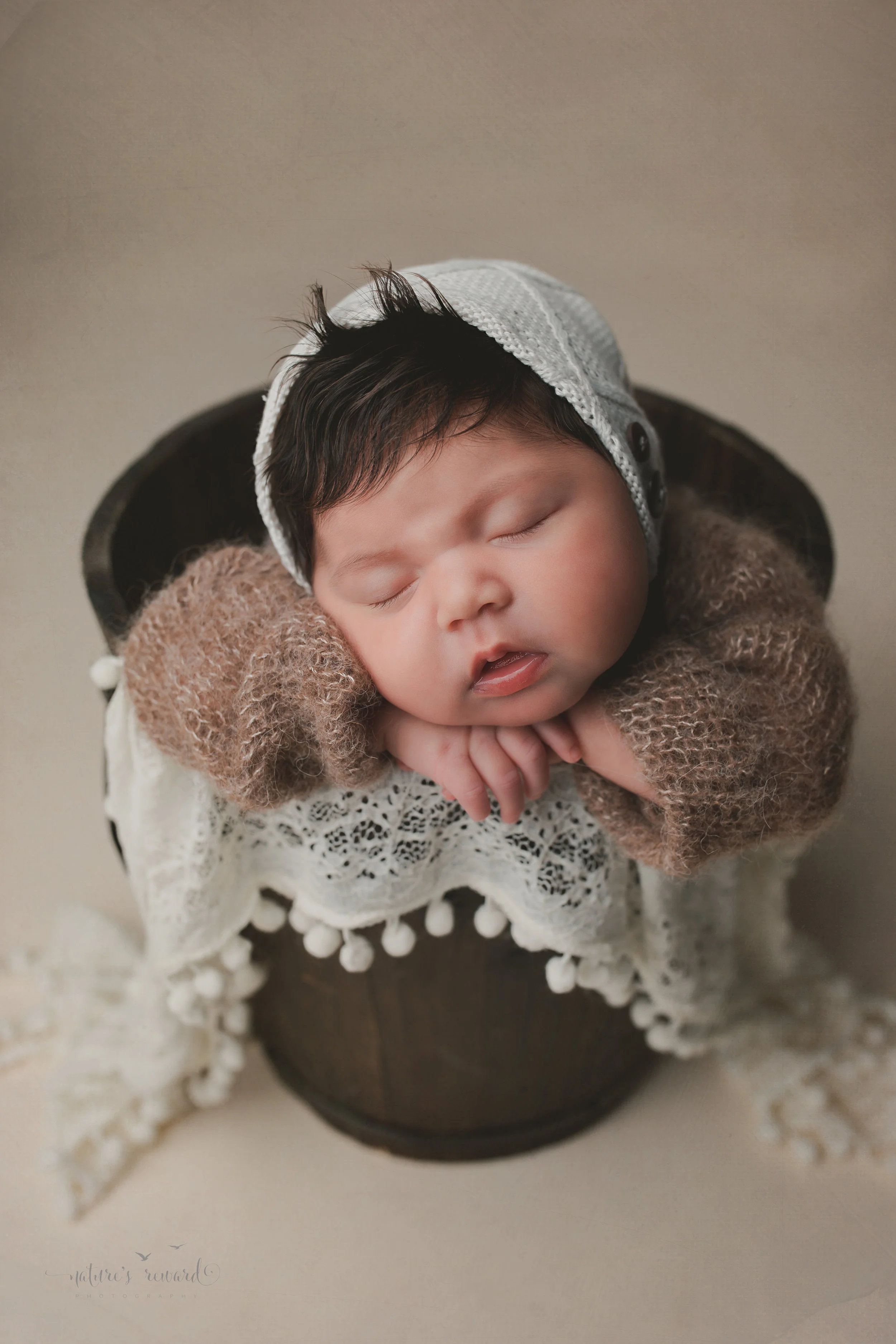 Newborn baby girl in a bucket on tan background, a portrait by Nature’s Reward Photography