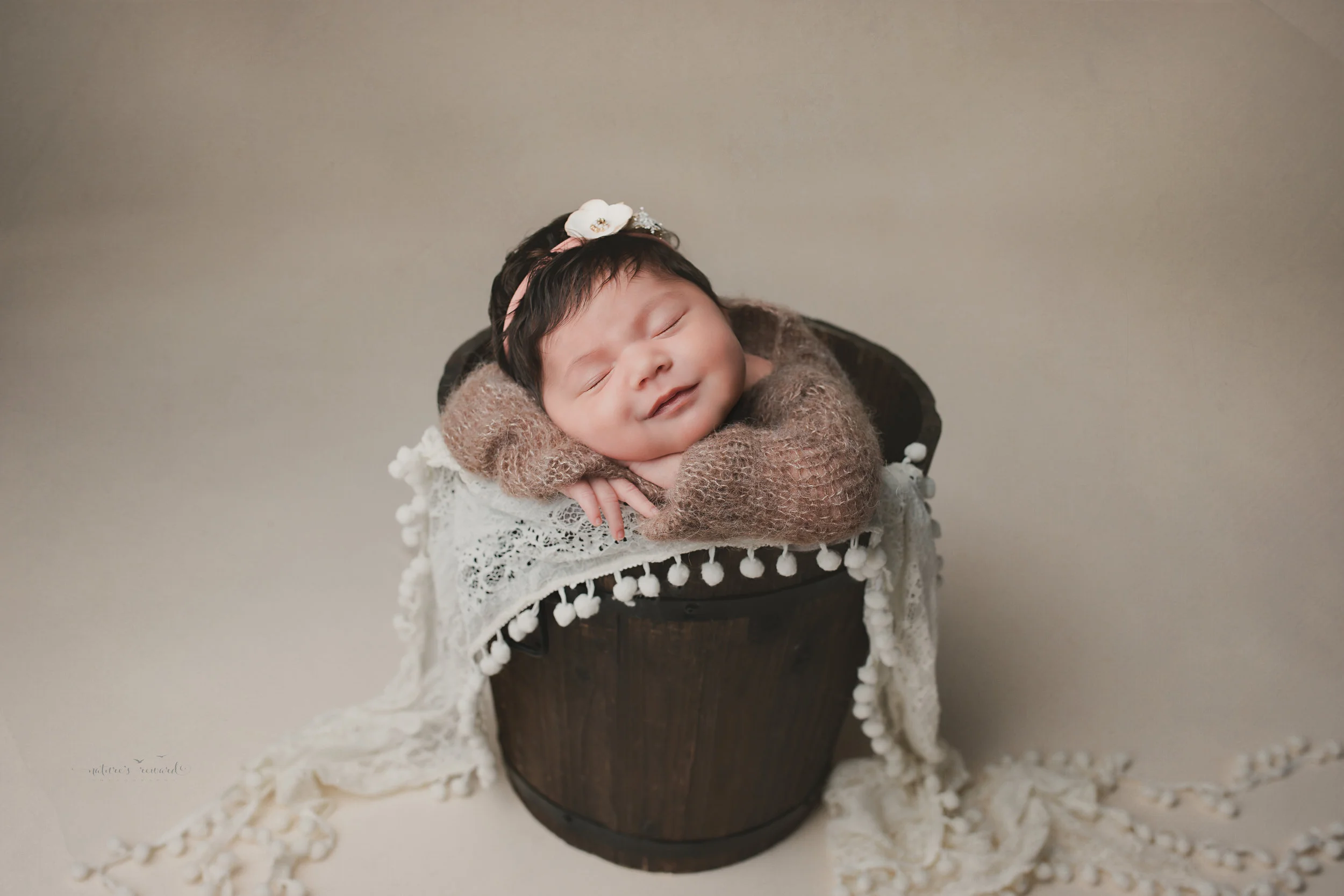 Newborn baby girl in a bucket on tan background, a portrait by Nature’s Reward Photography