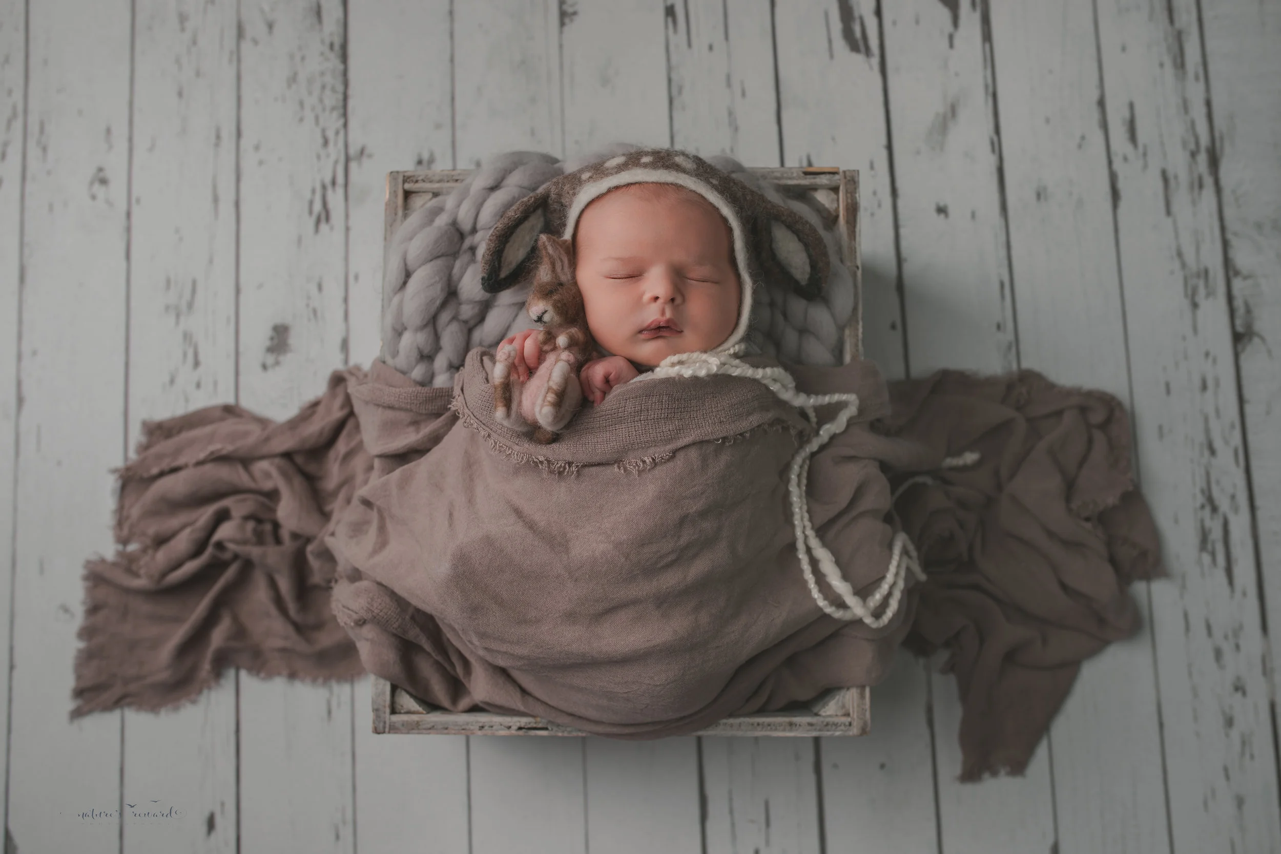 Newborn baby boy swaddled in white with brown scarf and wearing a deer bonnet, on a white wood floor, resting in a box, a portrait by Nature’s Reward Photography