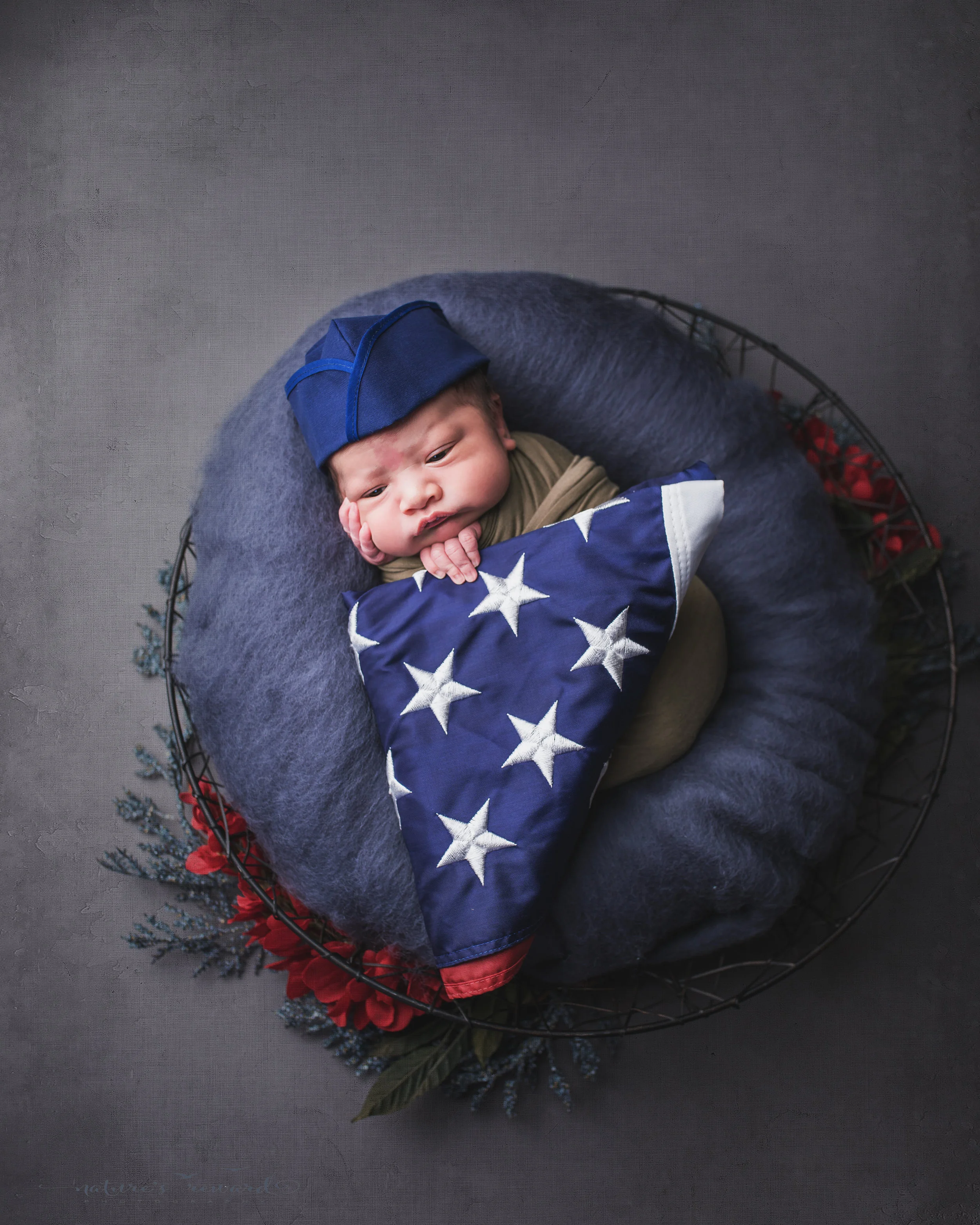 Wearing a service cap and holding an American flag, a swaddled newborn baby boy, a portrait by Nature’s Reward Photography