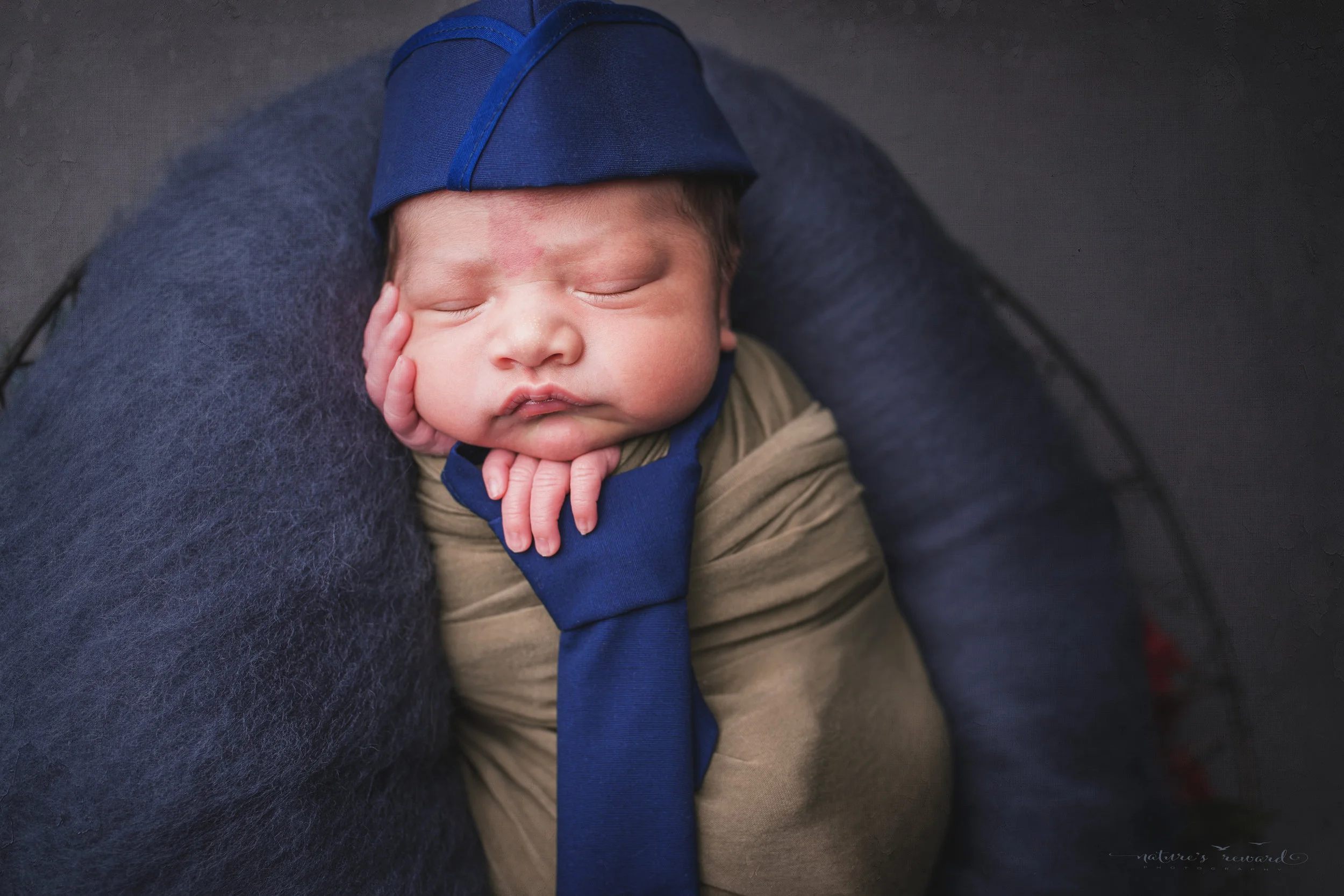 Wearing a service cap and holding an American flag, a swaddled newborn baby boy, a portrait by Nature’s Reward Photography
