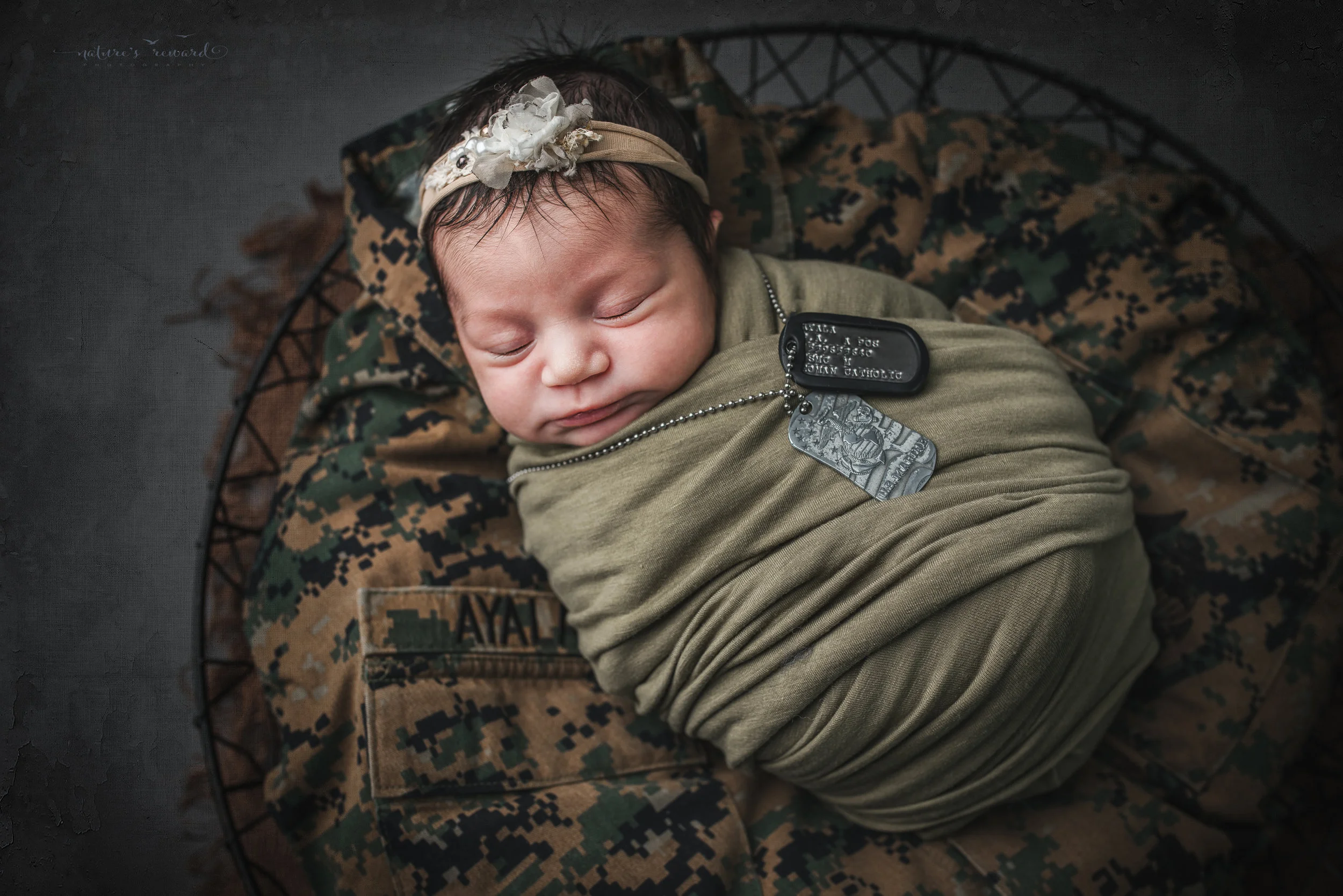 Newborn baby girl swaddled in army green laying on her father’s service uniform with his dog tags, a portrait by Nature’s Reward Photography