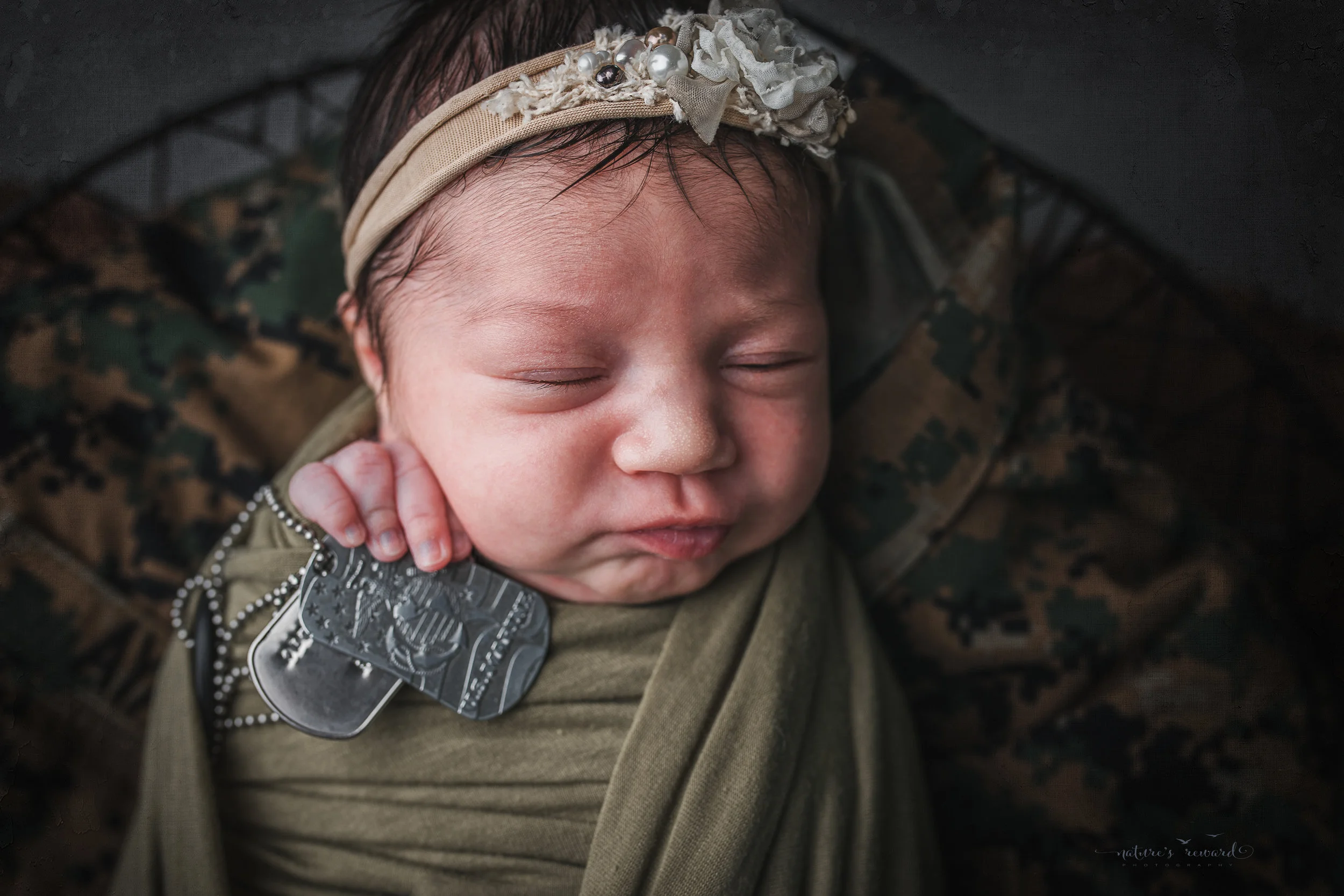 Newborn baby girl swaddled in army green laying on her father’s service uniform with his dog tags, a portrait by Nature’s Reward Photography