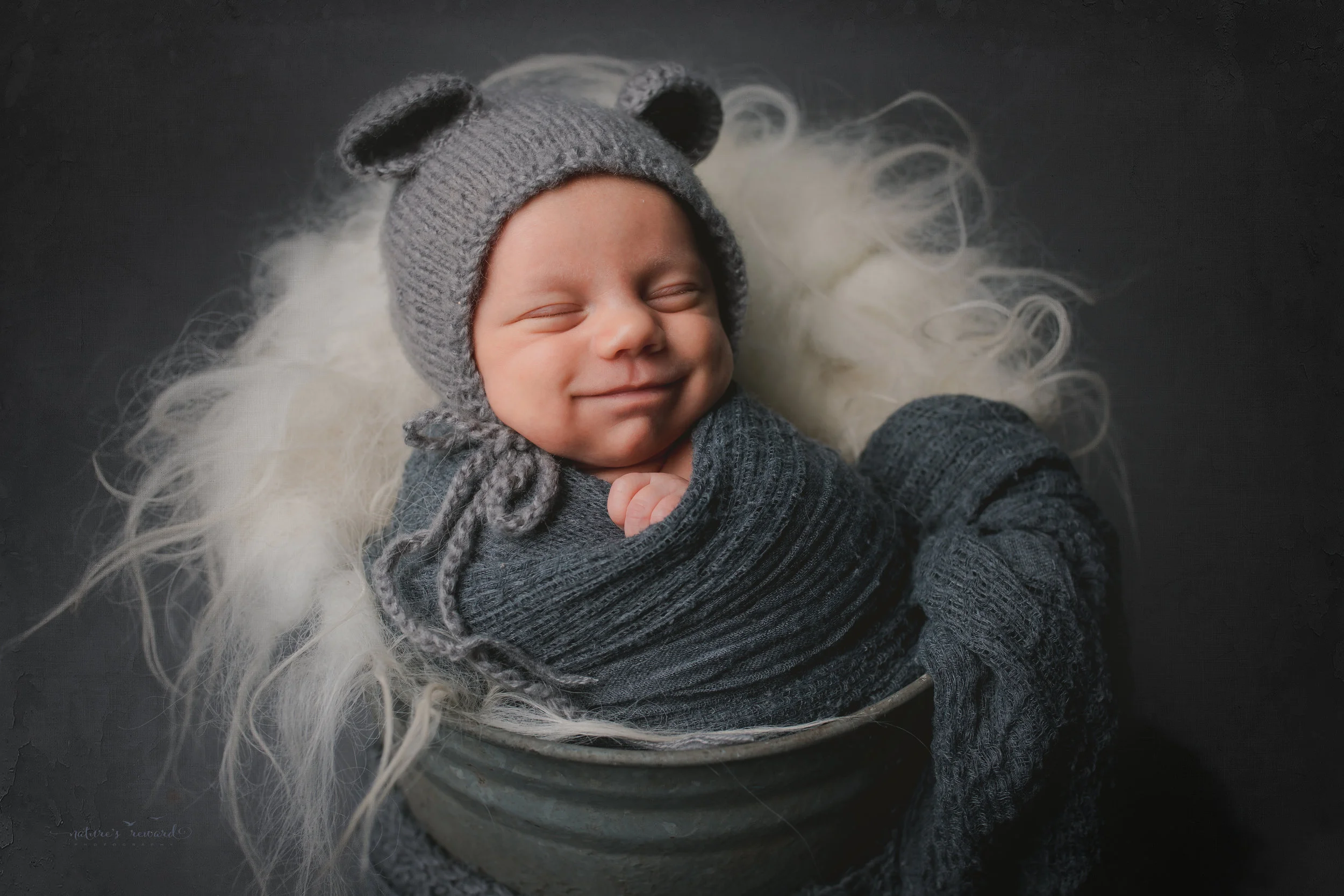 A baby boy in a bucket wearing a bear bonnet , smiling- a portrait by Nature's Reward Photography