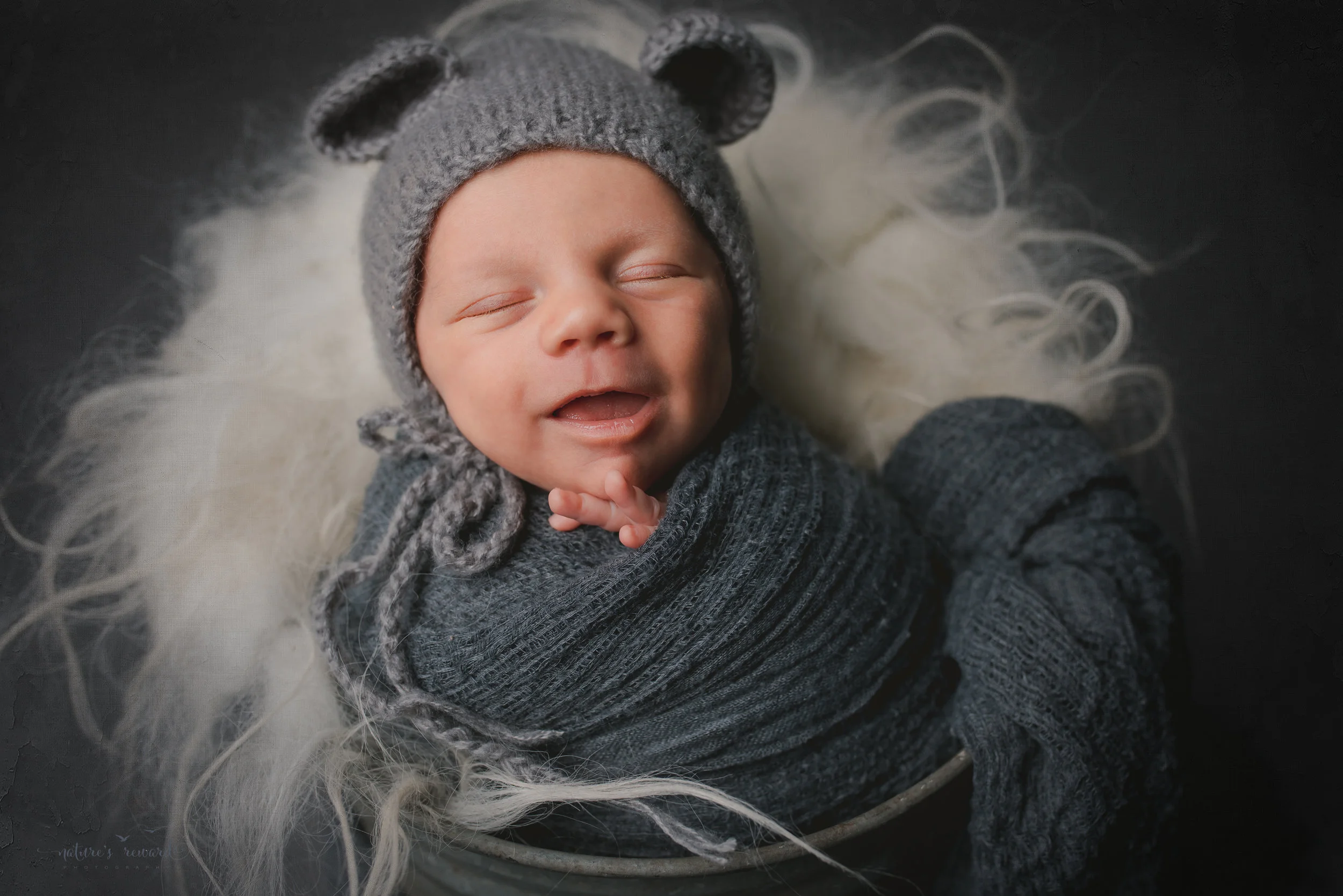 A baby boy in a bucket wearing a bear bonnet , smiling- a portrait by Nature's Reward Photography