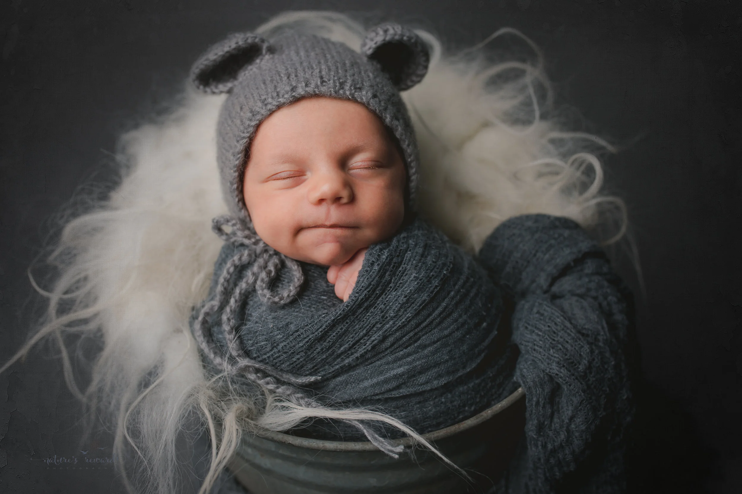 A baby boy in a bucket wearing a bear bonnet , grinning- a portrait by Nature's Reward Photography