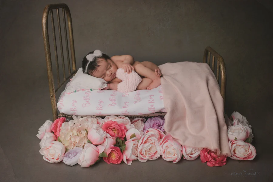 Newborn baby girl on a bed with her name written in the sheet surround by flowers in this portrait by Nature’s Reward Photography