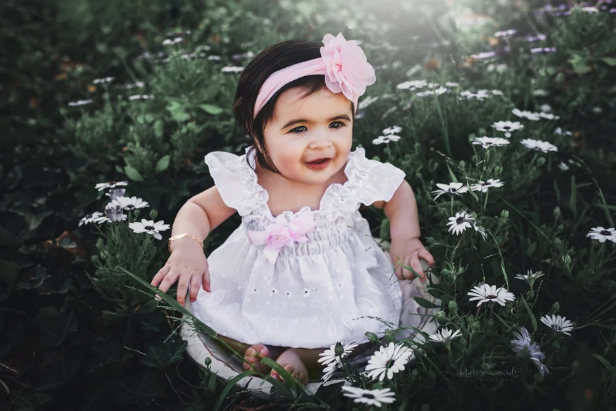 Sweet 6 month old baby girl in a summer white dress in the spring flower garden a portrait by Nature’s Reward Photography