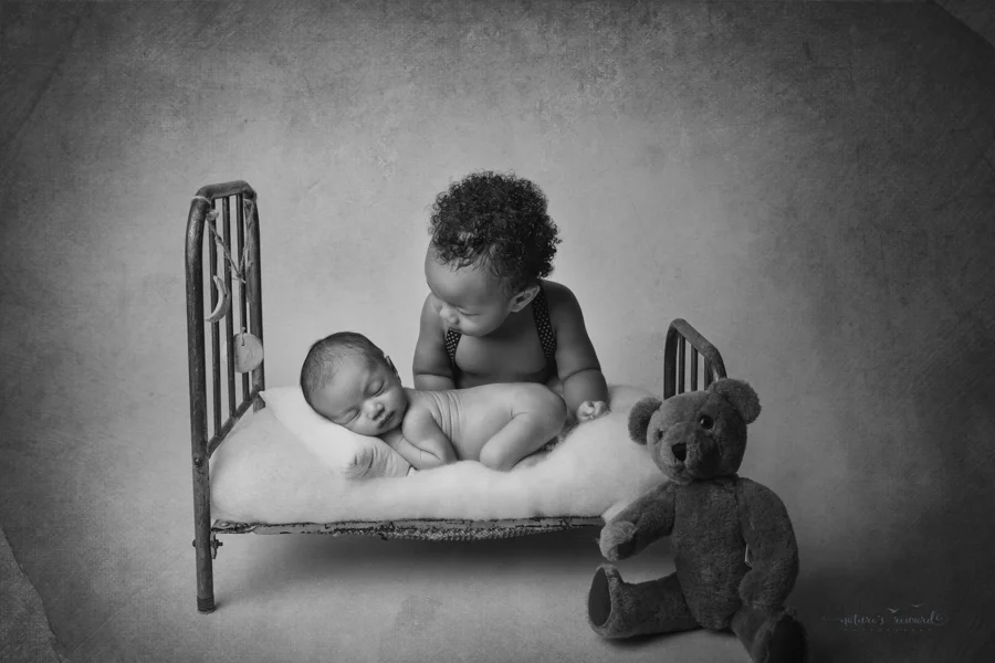 Black and white portrait of newborn baby boy laying in a bed in a bum up pose while his big brother looks lovingly at him in a newborn portrait by Nature’s Reward Photography