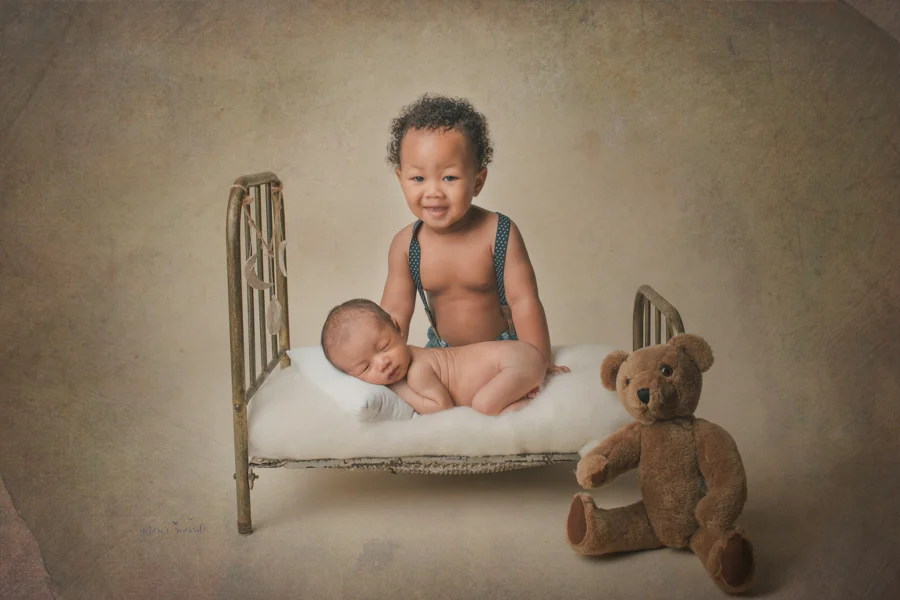 Newborn baby brother laying on a bed and his young big brother smiles excitedly in a newborn portrait by Nature’s Reward Photography