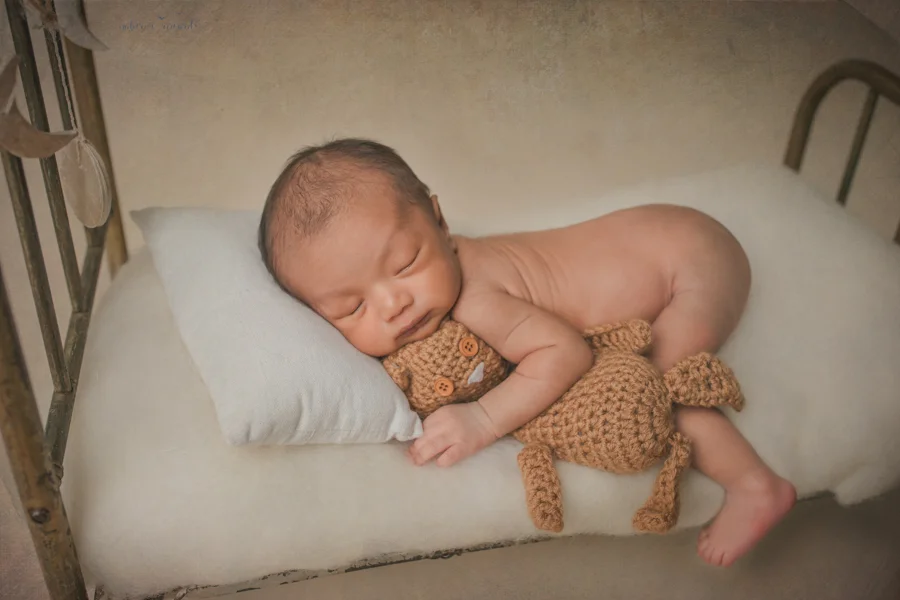 Newborn baby boy bum up with a leg dangling off the bed holding a teddy in a newborn portrait by Nature’s Reward Photography
