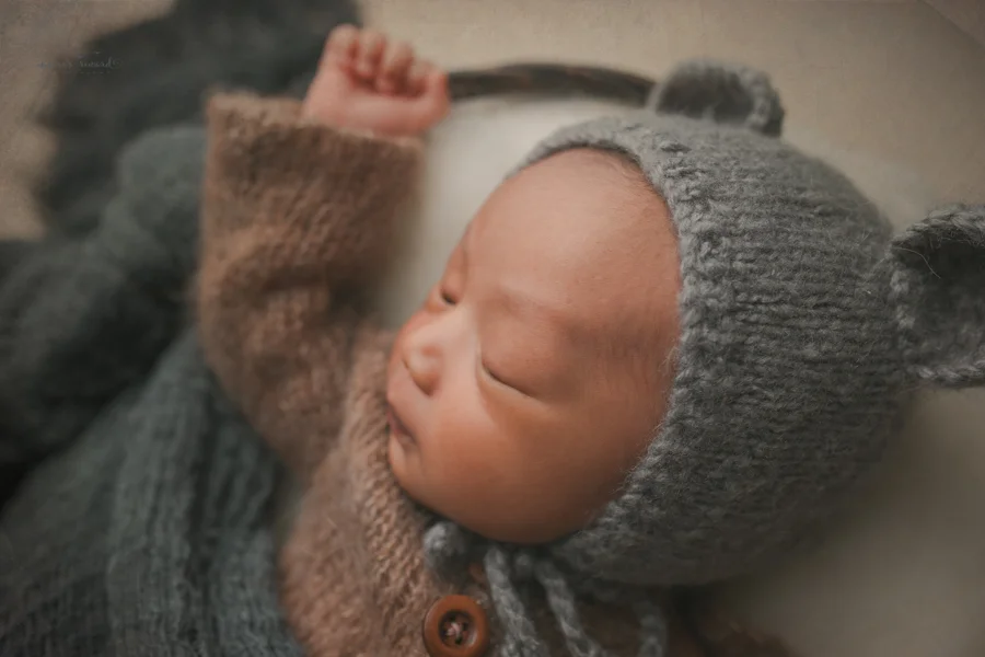 Newborn baby boy in a basket of fluff wearing a darling brown sleeper wearing a grey bear bonnet in a newborn portrait by Nature’s Reward Photography