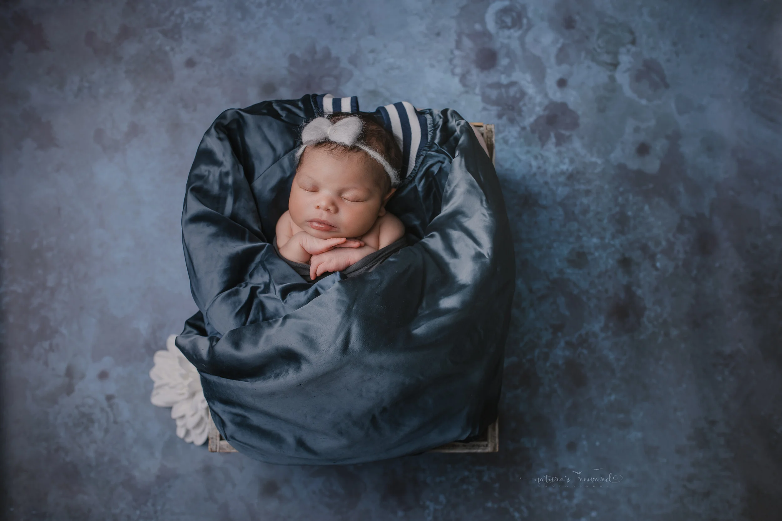 Newborn baby girl on a blue floral backdrop swaddled in her guardian angel grandfather’s jacket, a portrait by Nature’s Reward photography