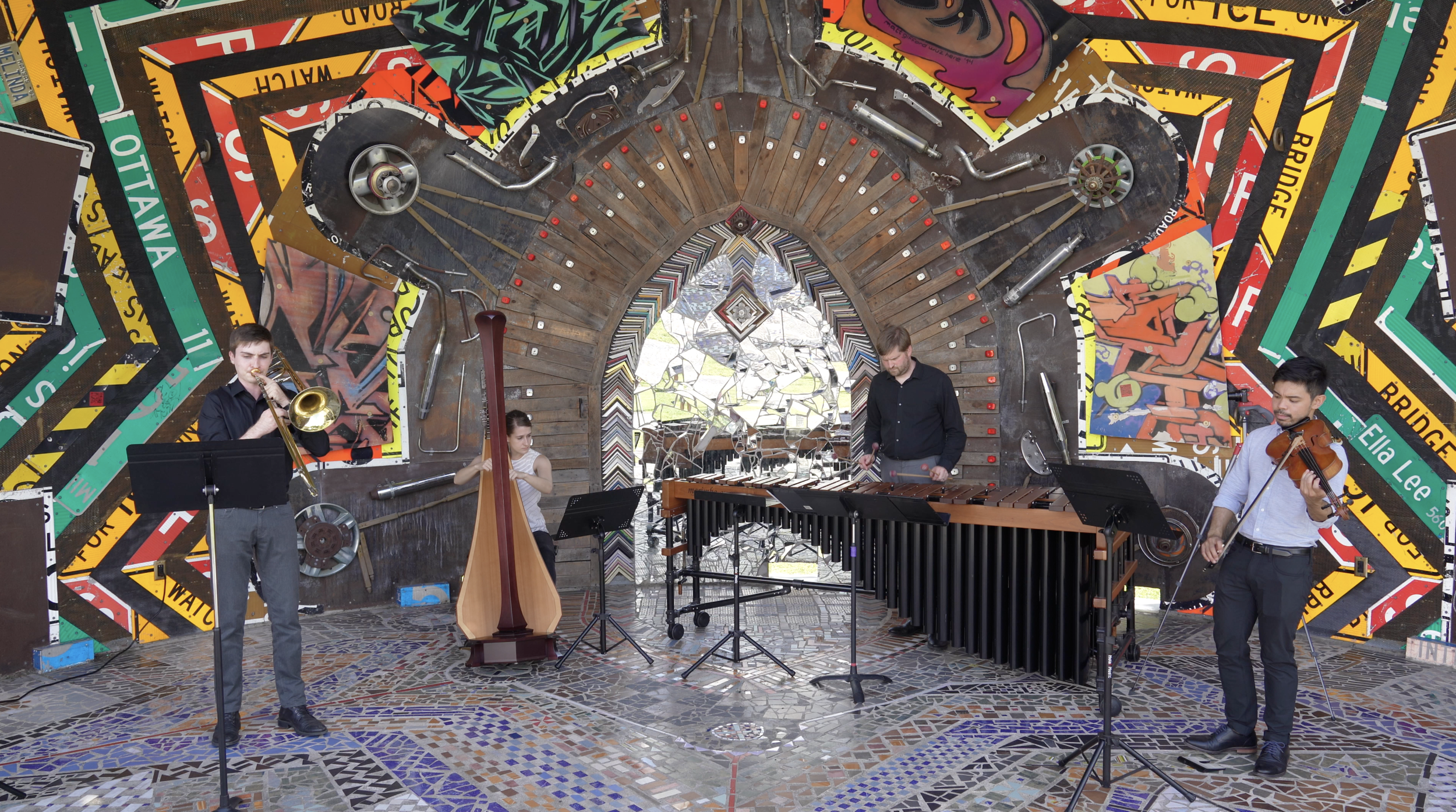 A trombonist, harpist, marimbaist, and violist performing outside in front of a colorful mosaic background, Smither Park.