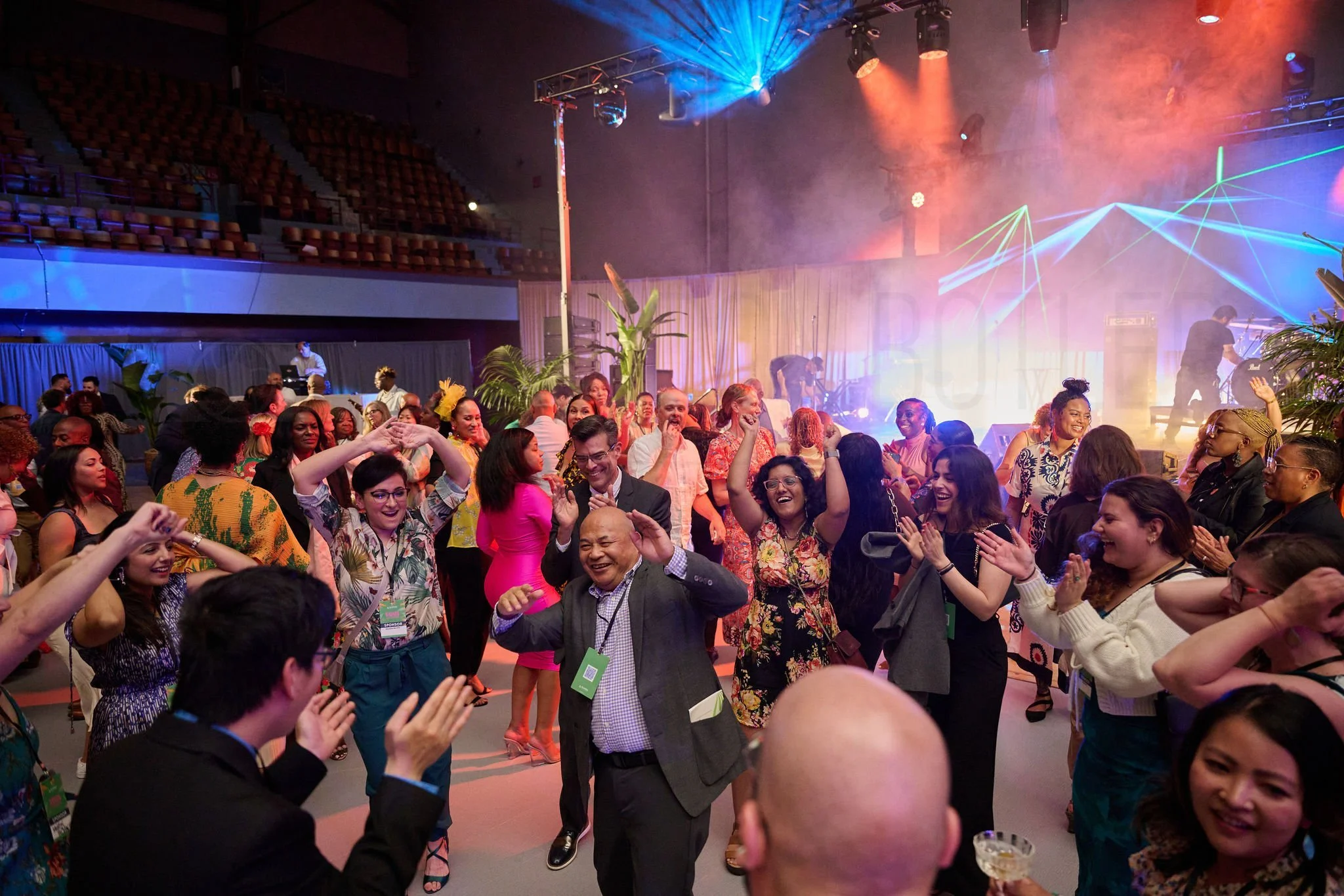 A lively indoor celebration with many smiling people dancing and enjoying themselves, illuminated by colorful stage lighting and laser effects.