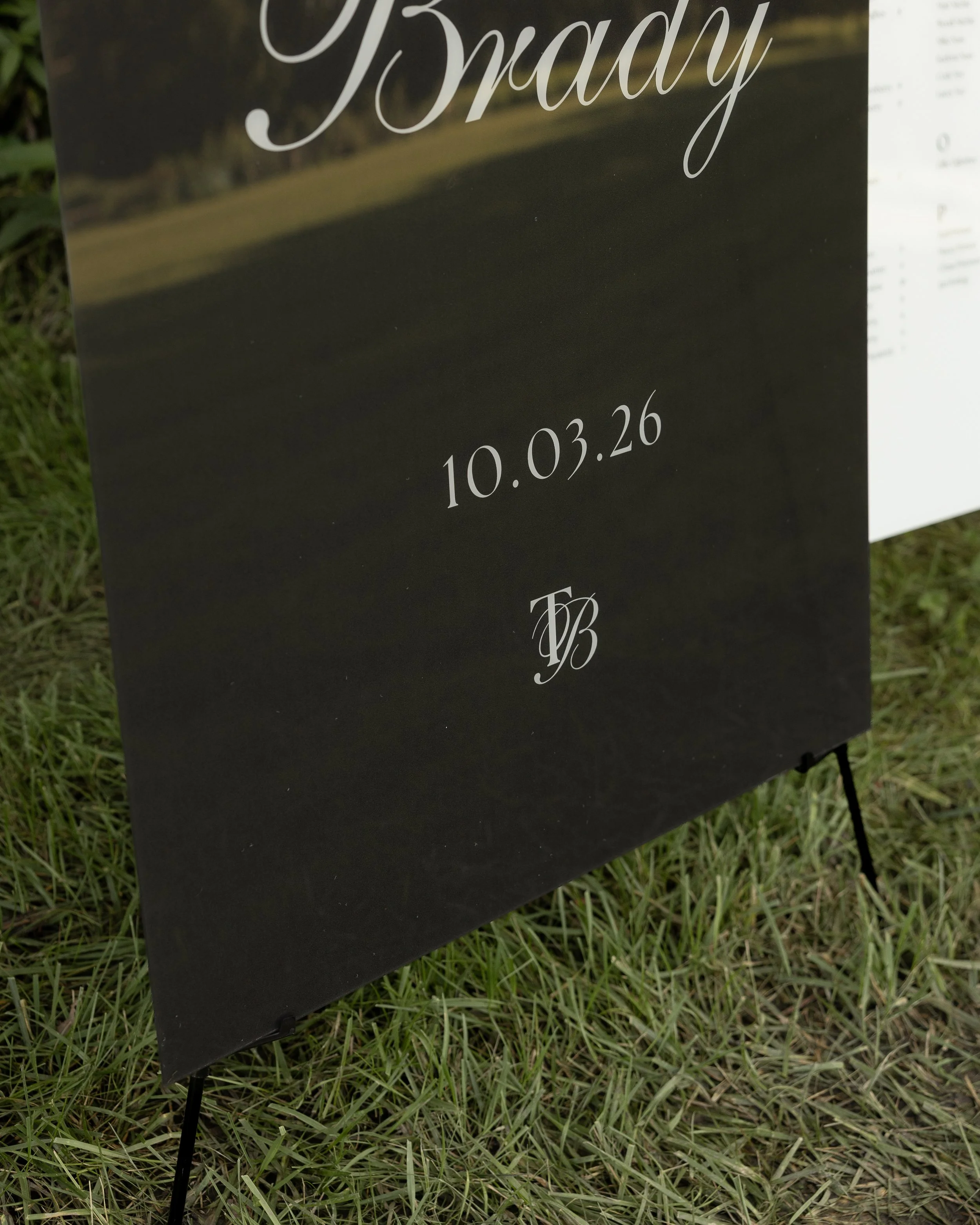 Close-up of a black sign with the word 'Brady' in white cursive font and the date 10.03.26 below, standing on grass with a blurred background.