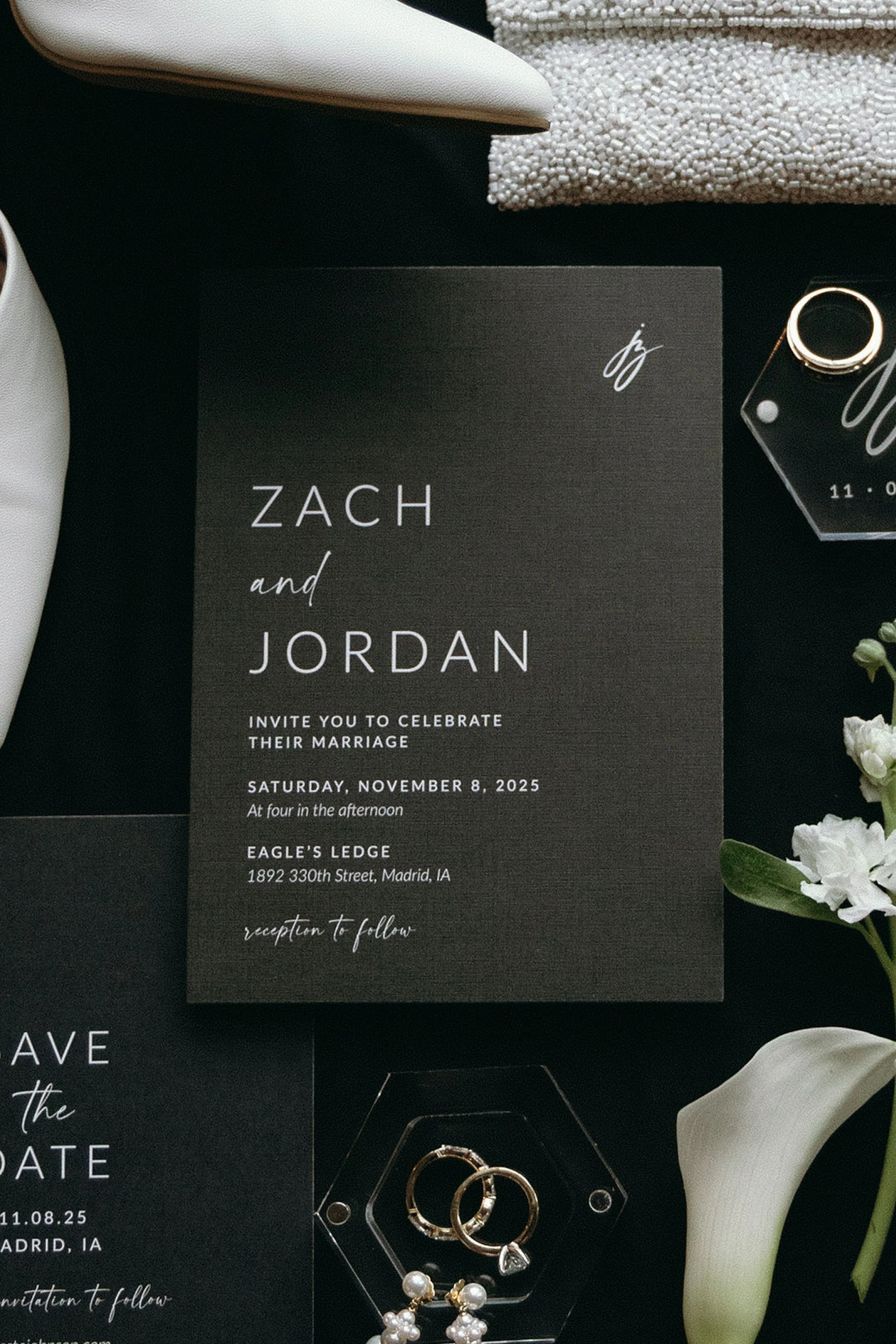 Wedding invitation card for Zach and Jordan with details of their marriage celebration on Saturday, November 8, 2025, at Eagle's Ledge in Madrid, IA, surrounded by jewelry, flowers, and decorative items for the event.