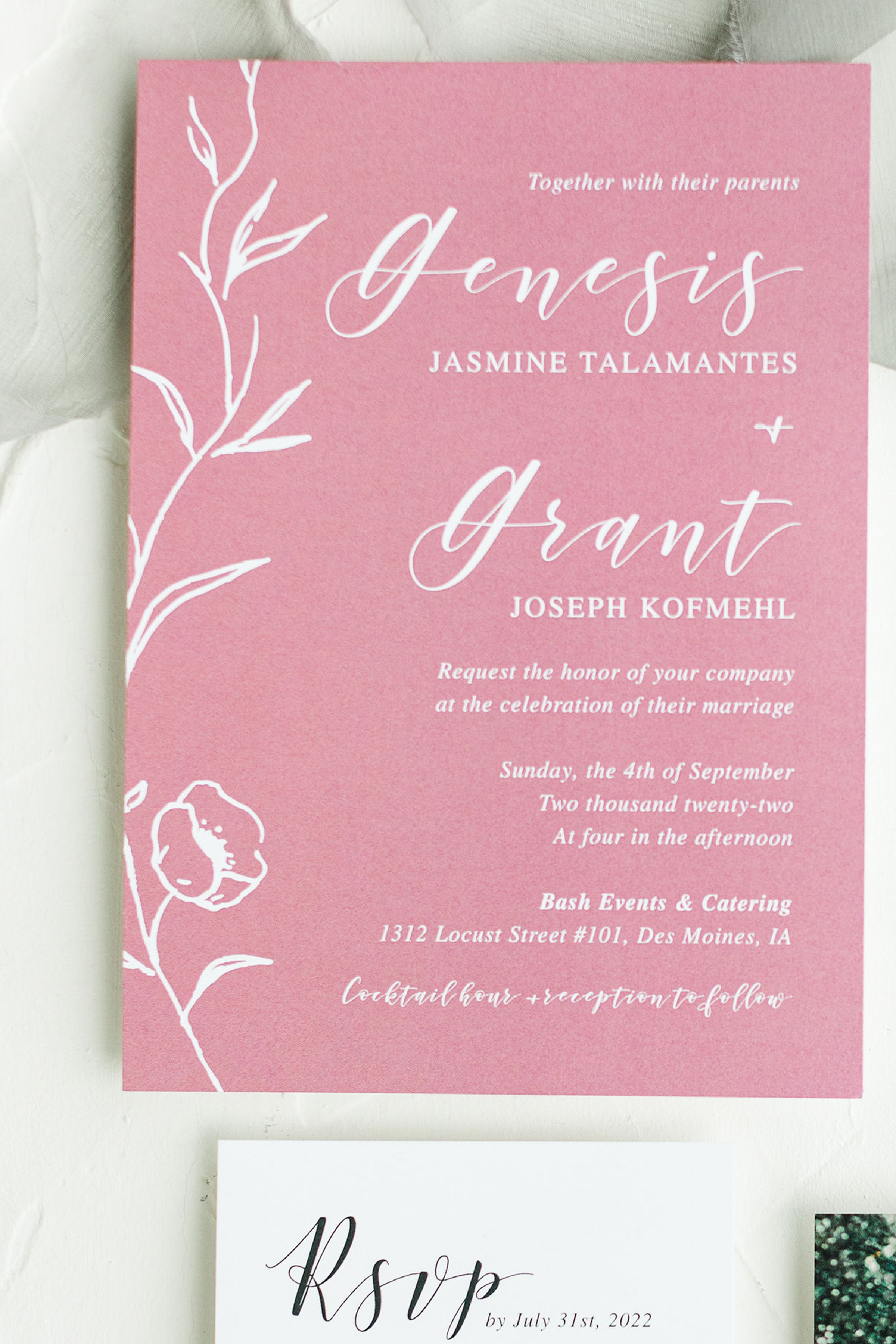 Close-up photograph of a pink wedding or event invitation card with floral design and elegant cursive and serif text, resting on a white fabric surface.