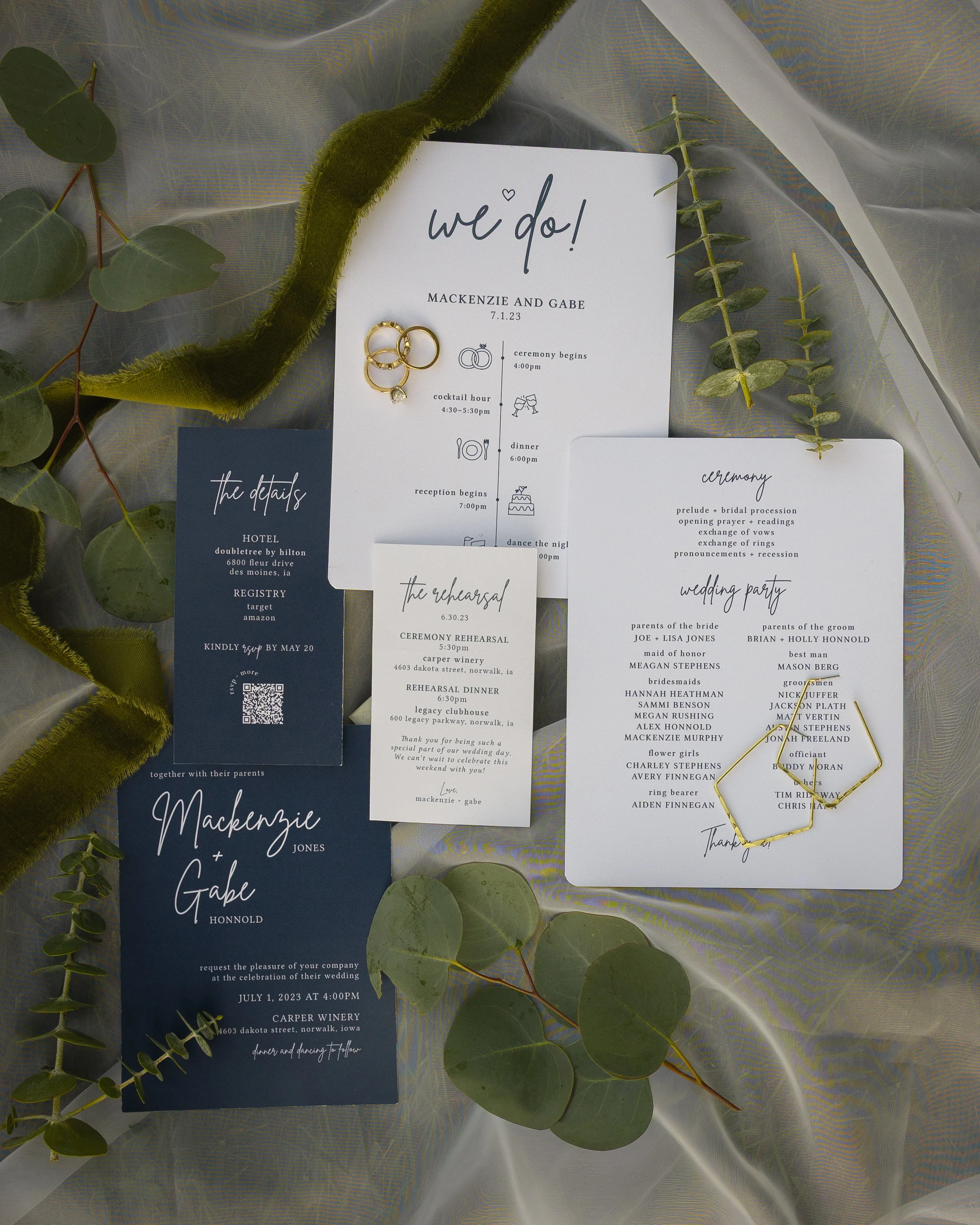 A wedding invitation suite with details about the ceremony, reception, and rehearsal dinner, accompanied by a gold wedding band and a diamond ring on top. The suite is decorated with green foliage and set on a semi-transparent fabric background.