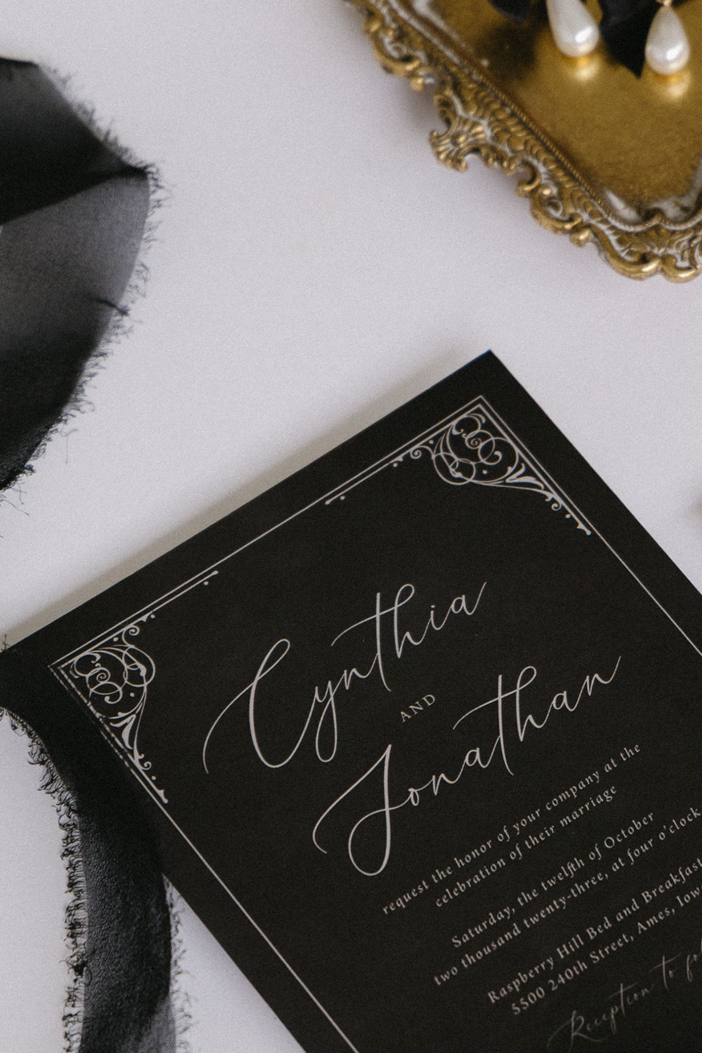 Close-up of a wedding invitation card with elegant cursive text and decorative borders, placed on a white surface with a black fabric and a gilded ornate box partially visible.