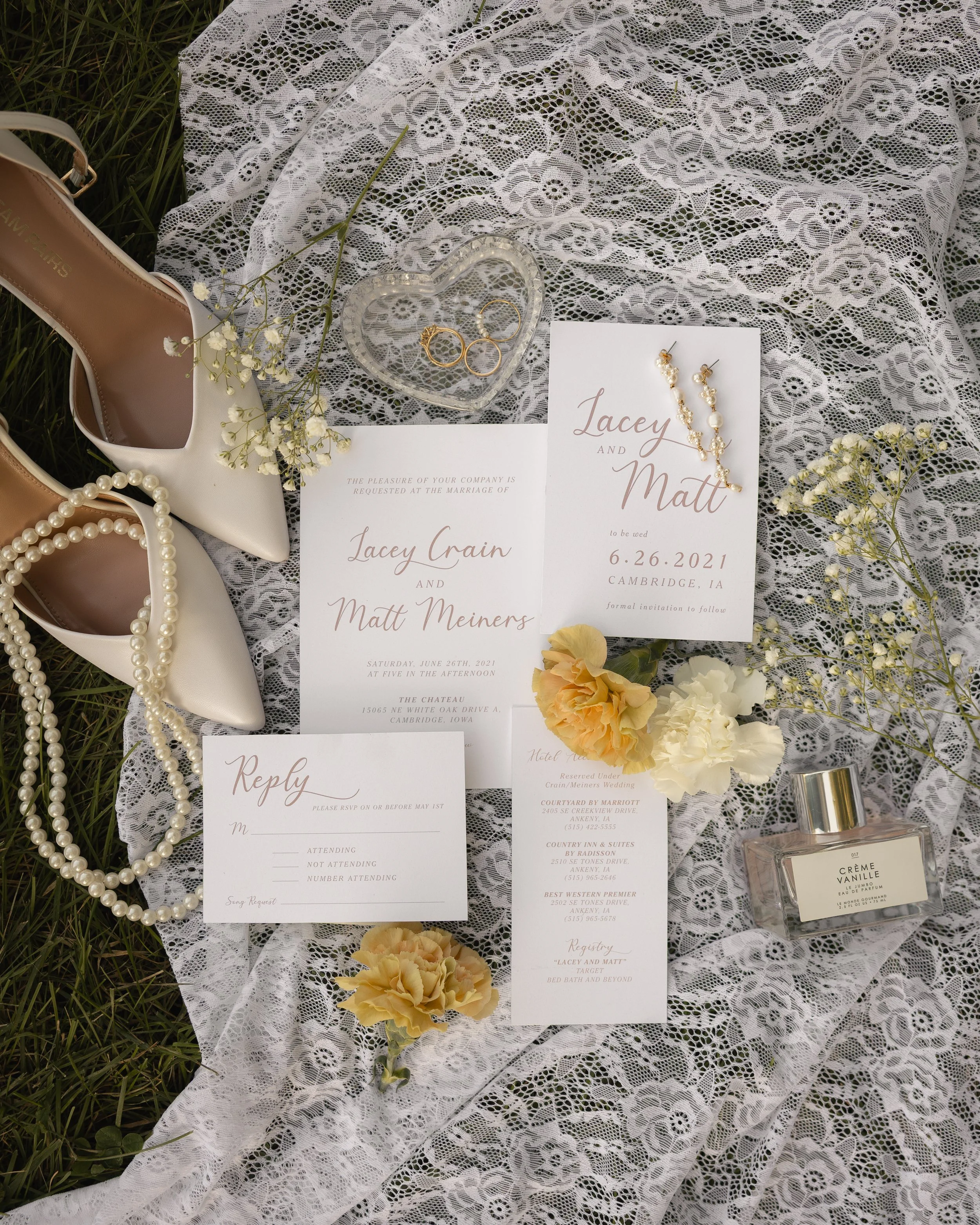 Wedding invitation on lace fabric with flowers, jewelry, perfume, and shoes around it.