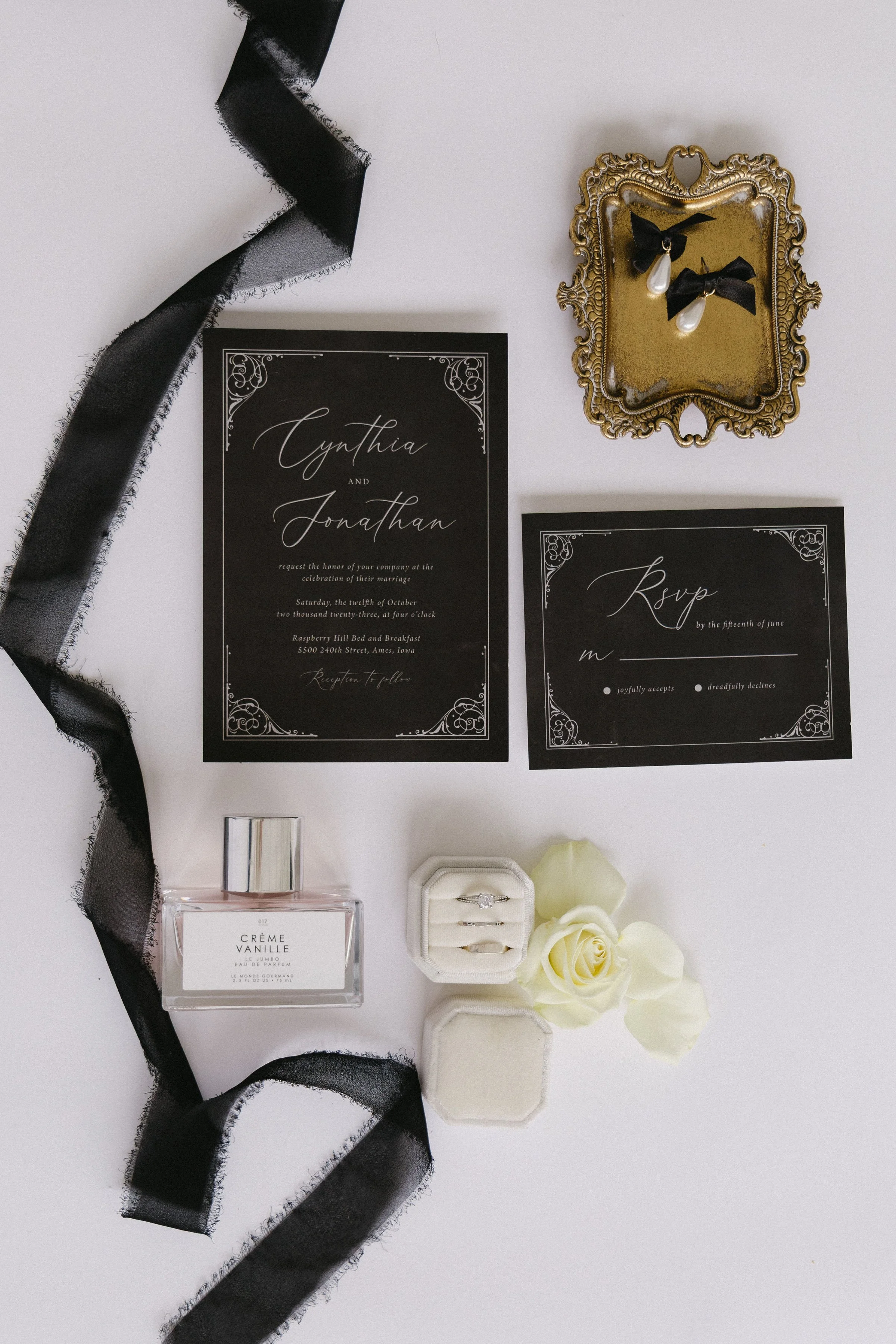 Invitation cards for a wedding with names Cynthia and Jonathan, a perfume bottle labeled Crème Vanille, a jewelry box with rings, cream-colored roses, and decorative accessories. Black ribbon is draped across the image.