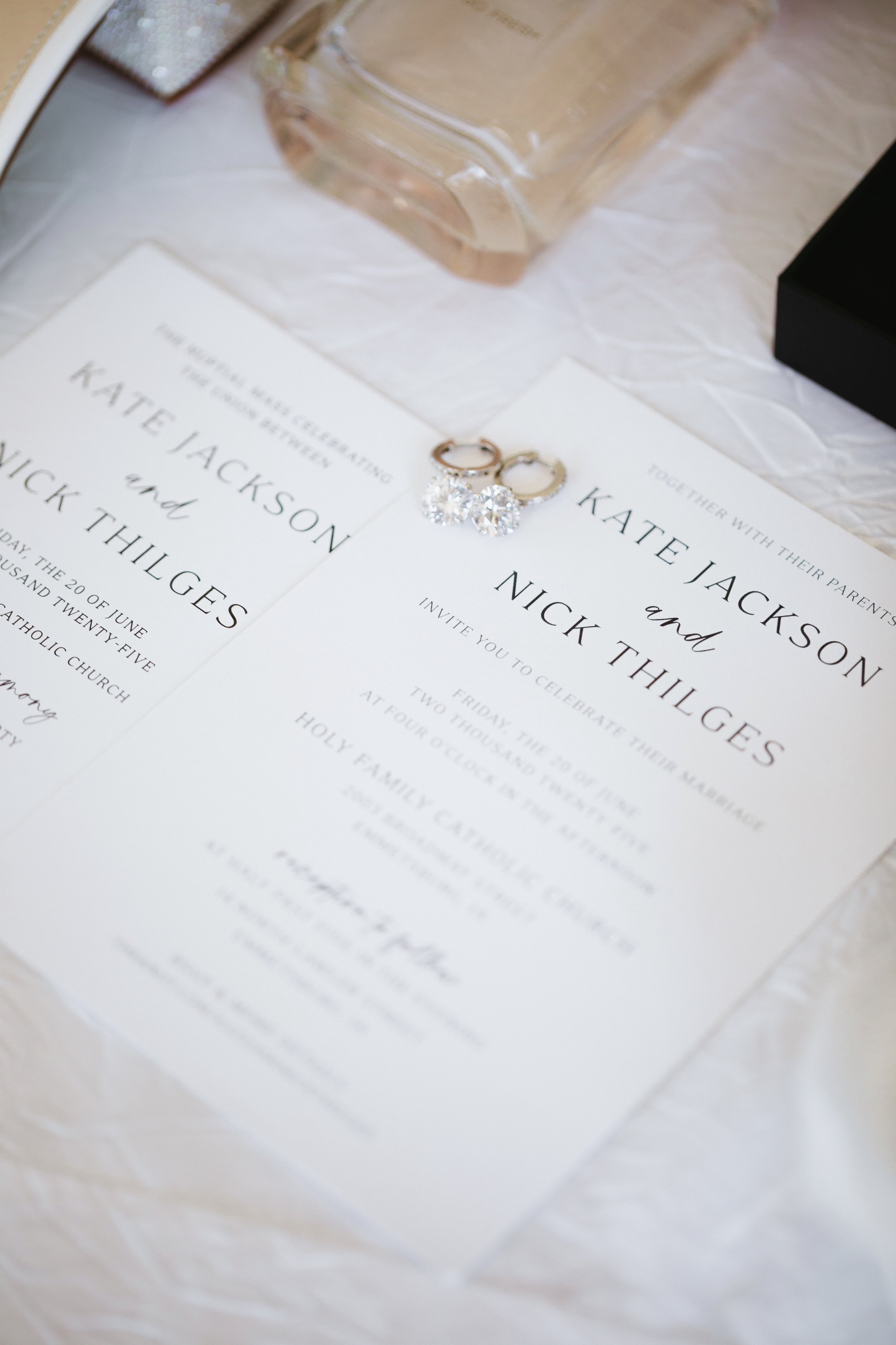 A wedding invitation with black text on white paper, with a pair of diamond earrings resting on top, and a clear perfume bottle and framed picture in the background.