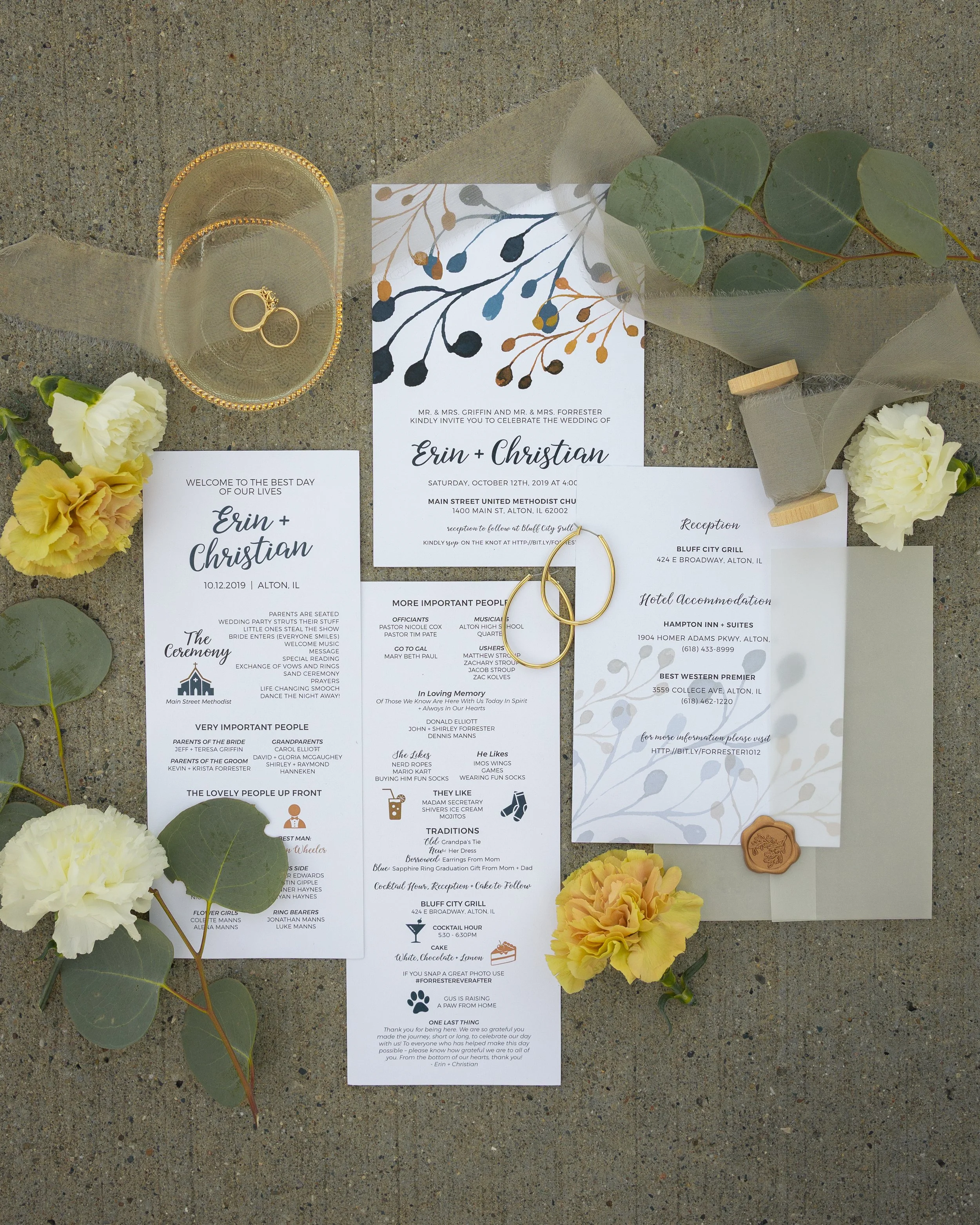 Wedding invitation, ceremony details, and reception information displayed on white paper, surrounded by white, yellow, and peach flowers, eucalyptus branches, a gold wedding ring, and a wax seal, all arranged on concrete.