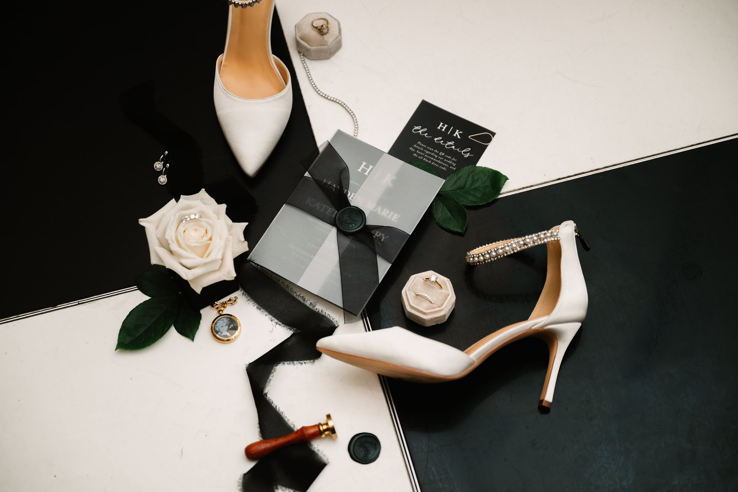 A collection of wedding accessories and jewelry including a white high heel shoe, a white rose with a ring, pearl earrings, a black ribbon with a seal, a small hexagonal box with a ring, a pearl-embellished clutch, and a wedding invitation on a black