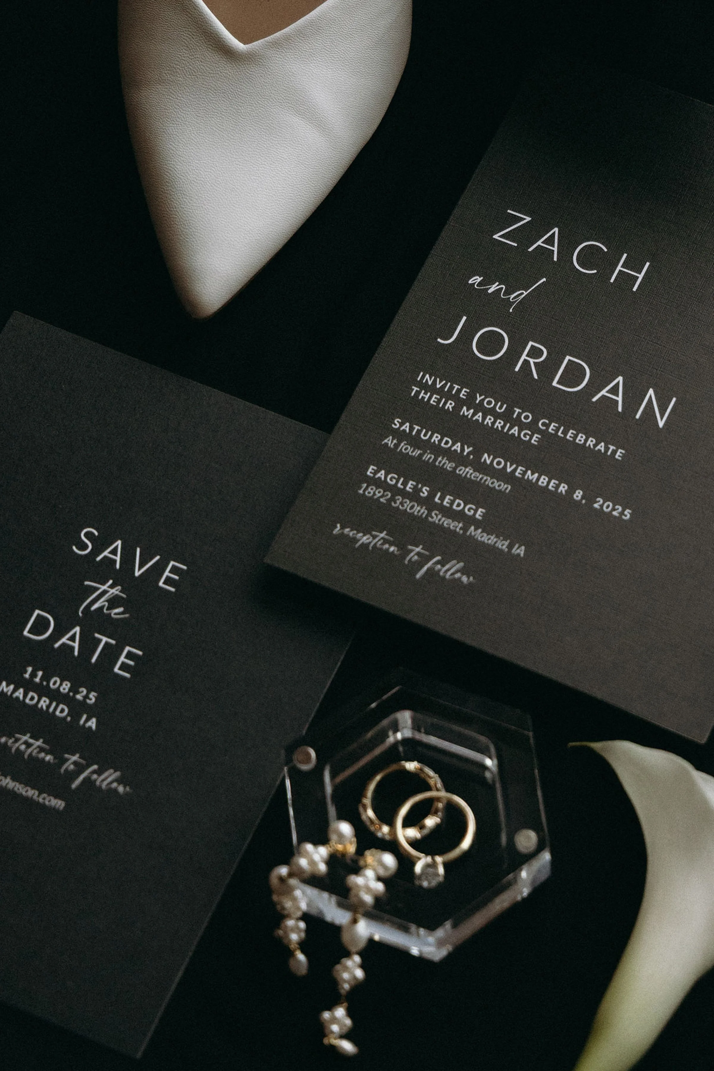 Invitation card for a wedding celebration of Zach and Jordan on November 8, 2025, with black background and white text, placed on a black surface next to jewelry including pearl earrings and rings.