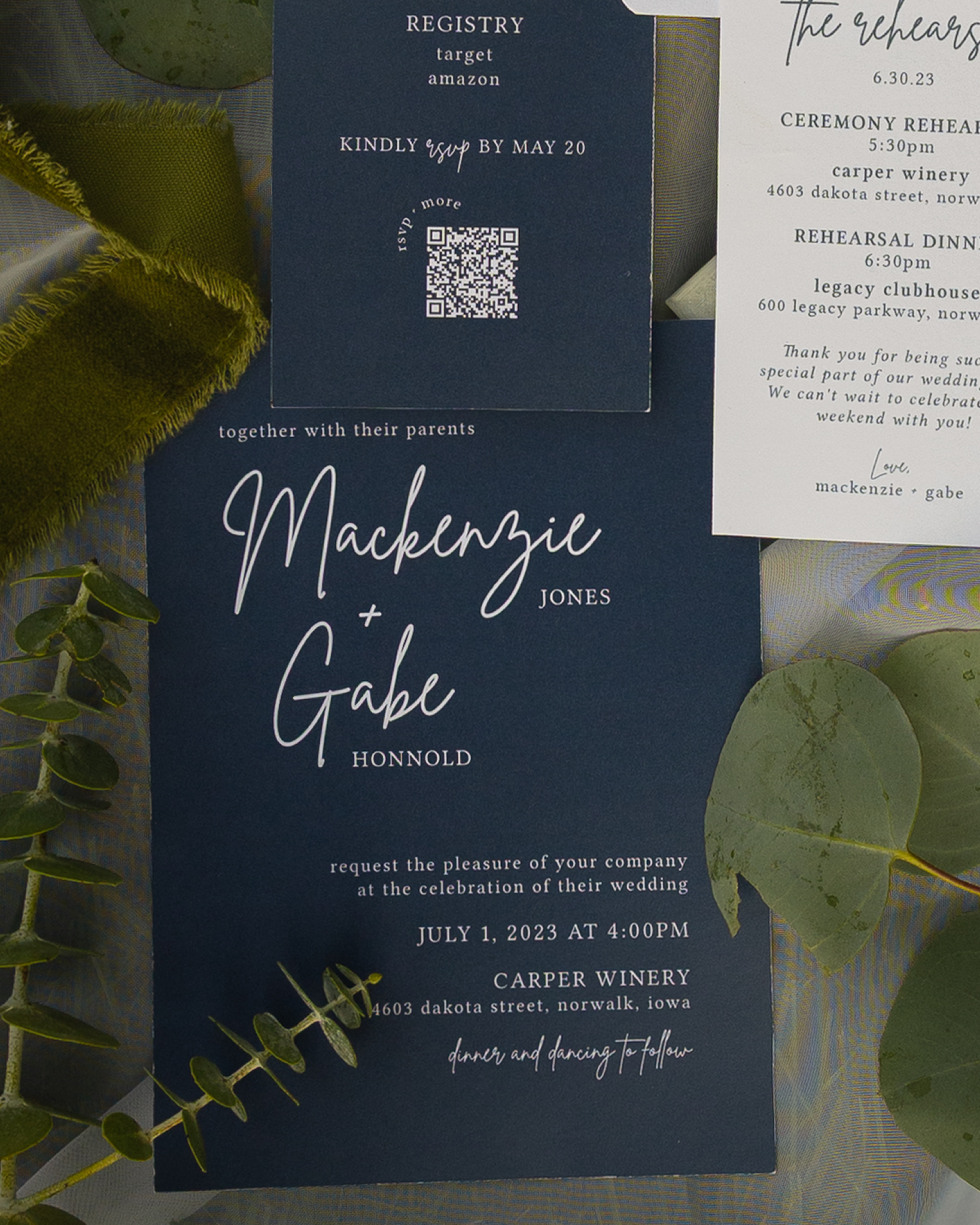 A wedding invitation with a dark blue background and white text. It includes details about a wedding registration, RSVP QR code, and event schedule at Carper Winery and Legacy Clubhouse in Norwalk, Iowa, with decorative green leaves around the invita