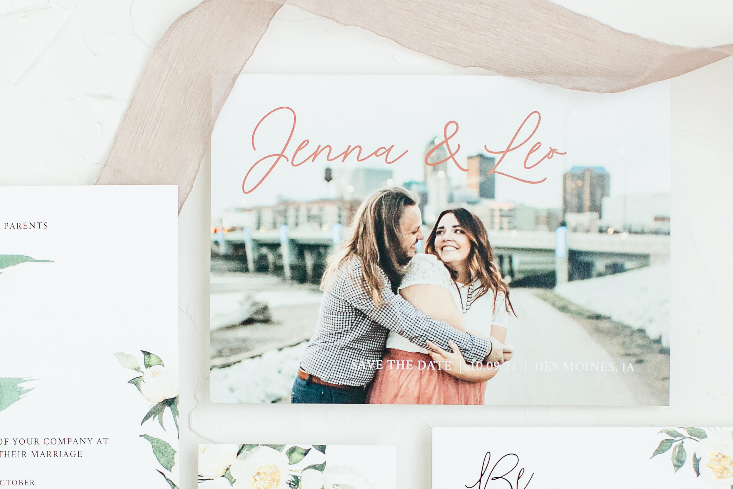 A wedding or engagement photo of two women hugging, with an urban cityscape in the background and a sign with text that reads 'Jenna & Leo'.